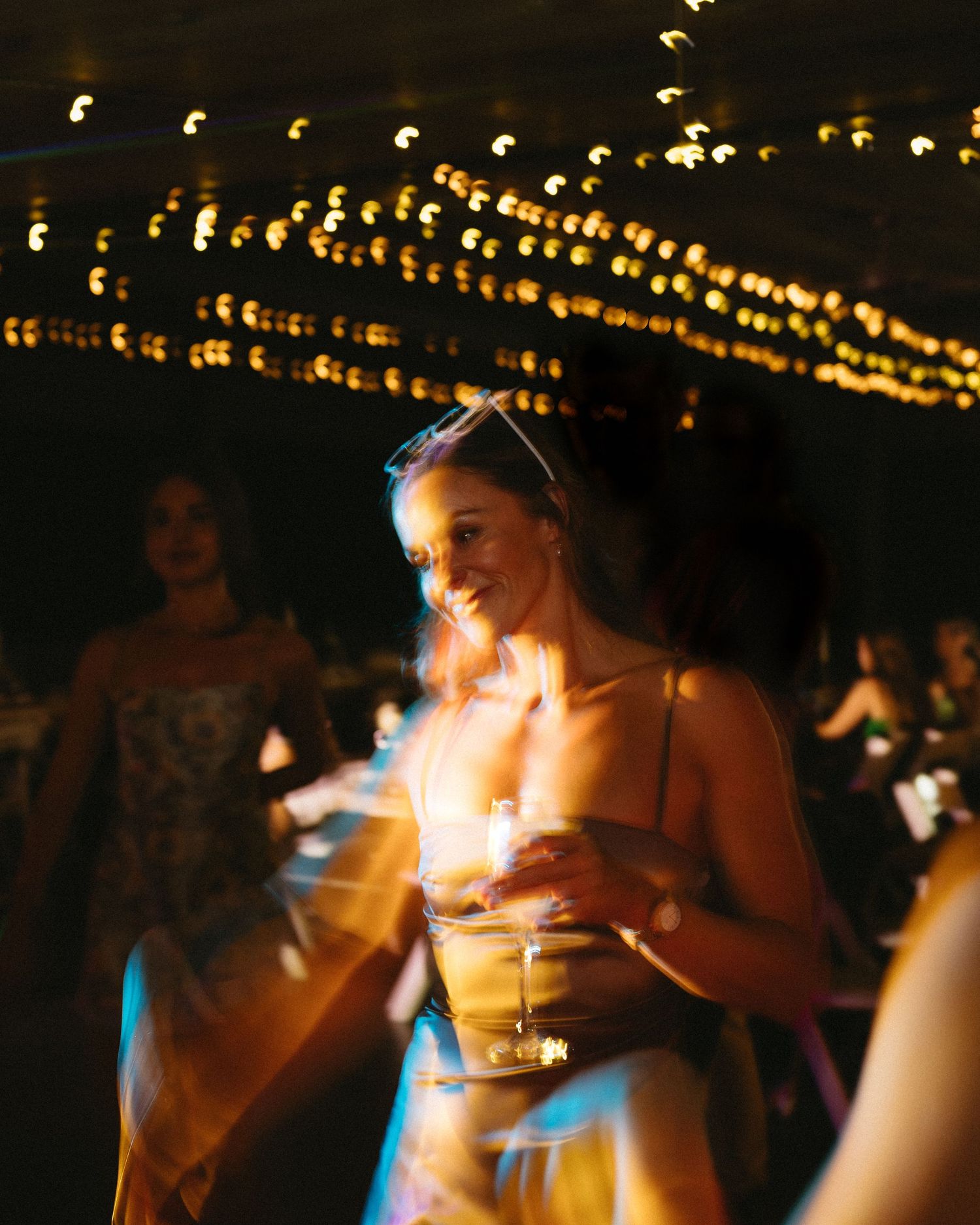 Wedding guest illuminated by warm string lights holds drink at reception.