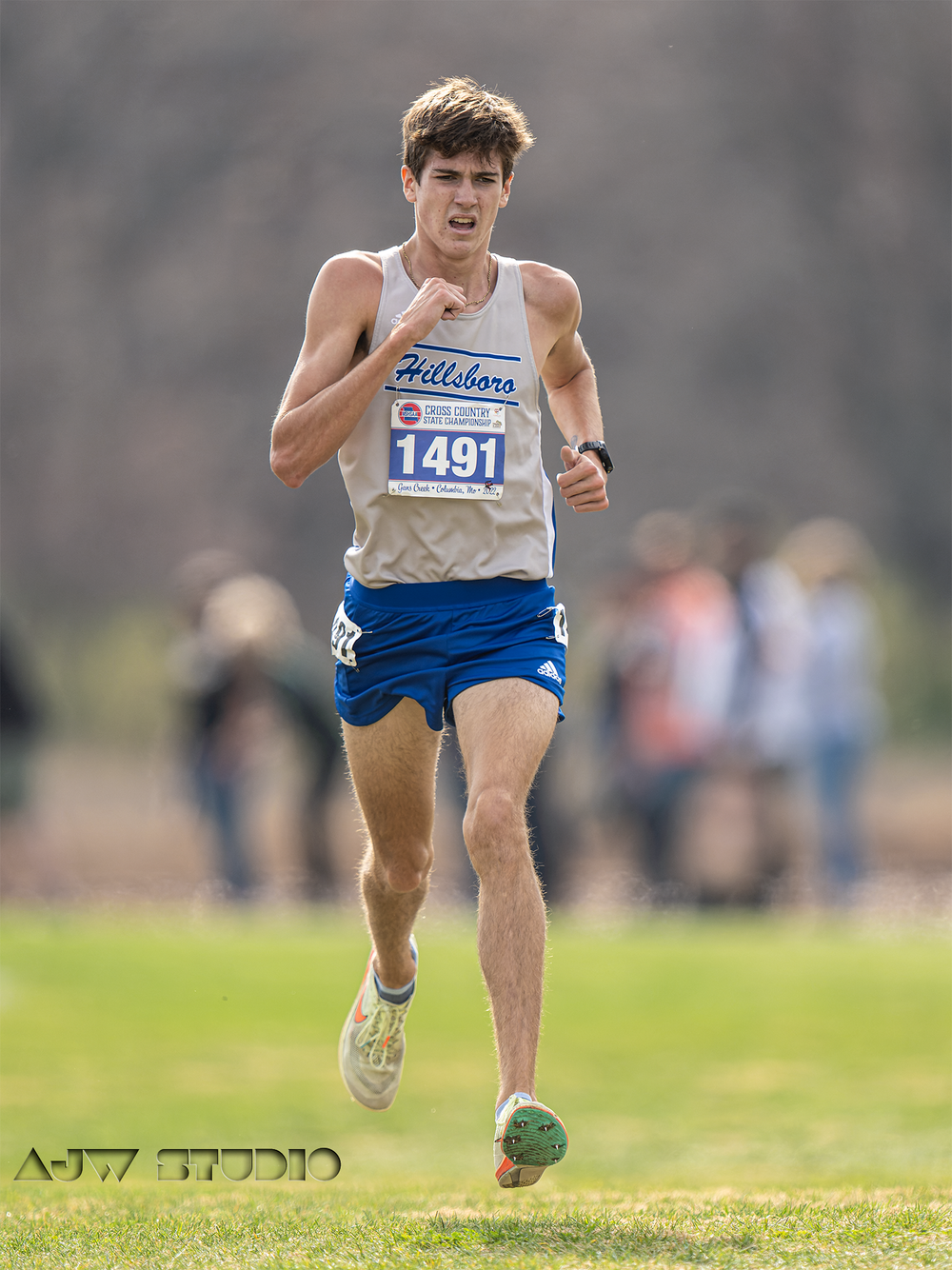 Cross country runner in white singlet and blue shorts races on grass course.