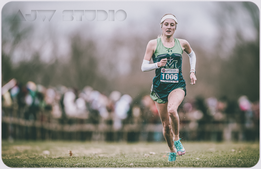 Cross country athlete in green uniform runs along a grass course during competition.