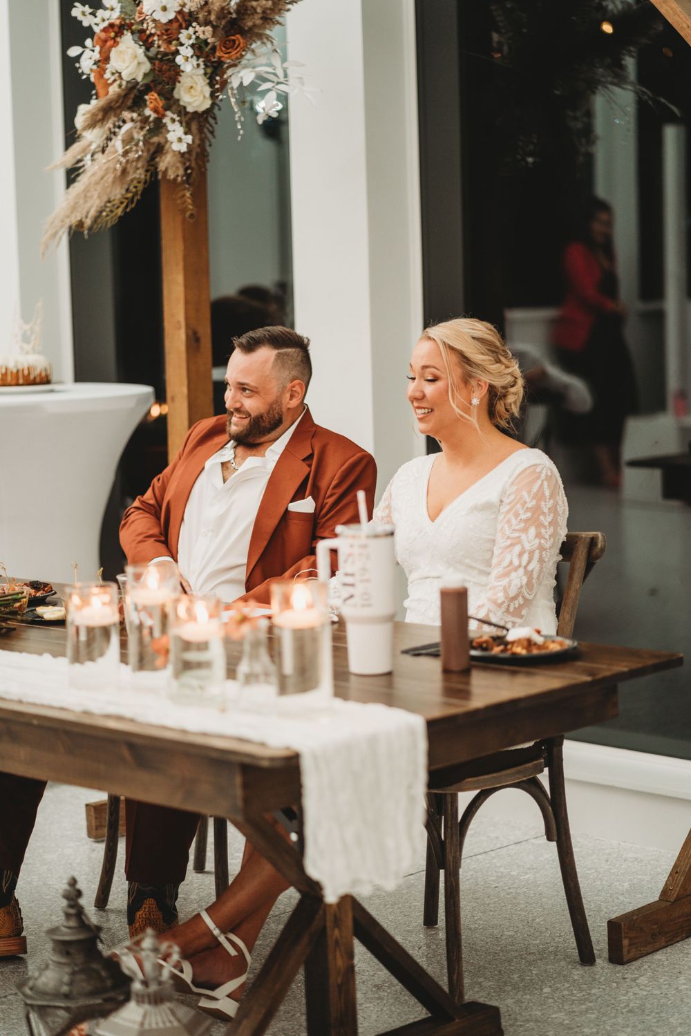 Couple seated at rustic reception table with boho floral arrangements and candlelight.