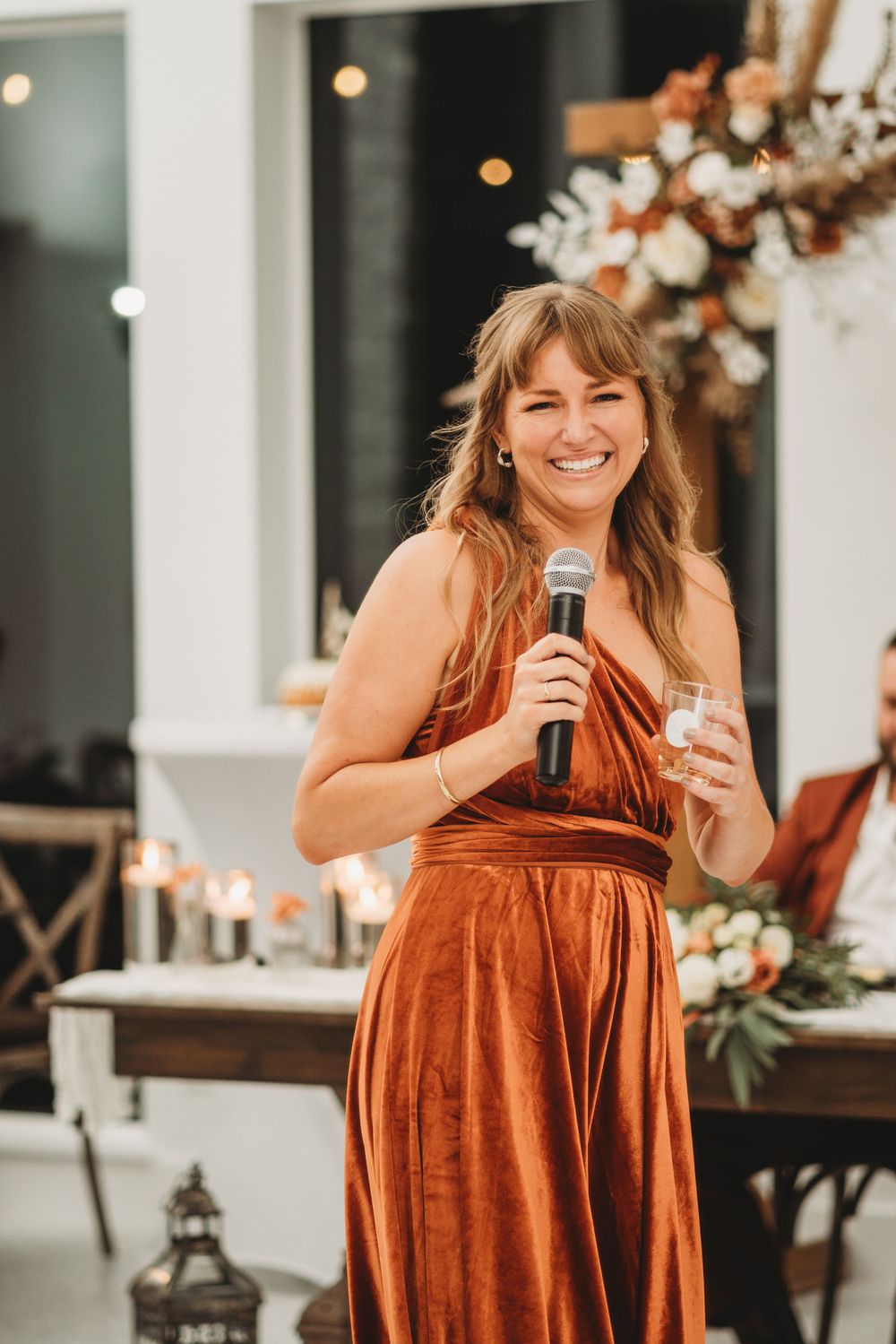 Wedding guest in a rust-colored dress delivers a speech at an elegant reception venue.