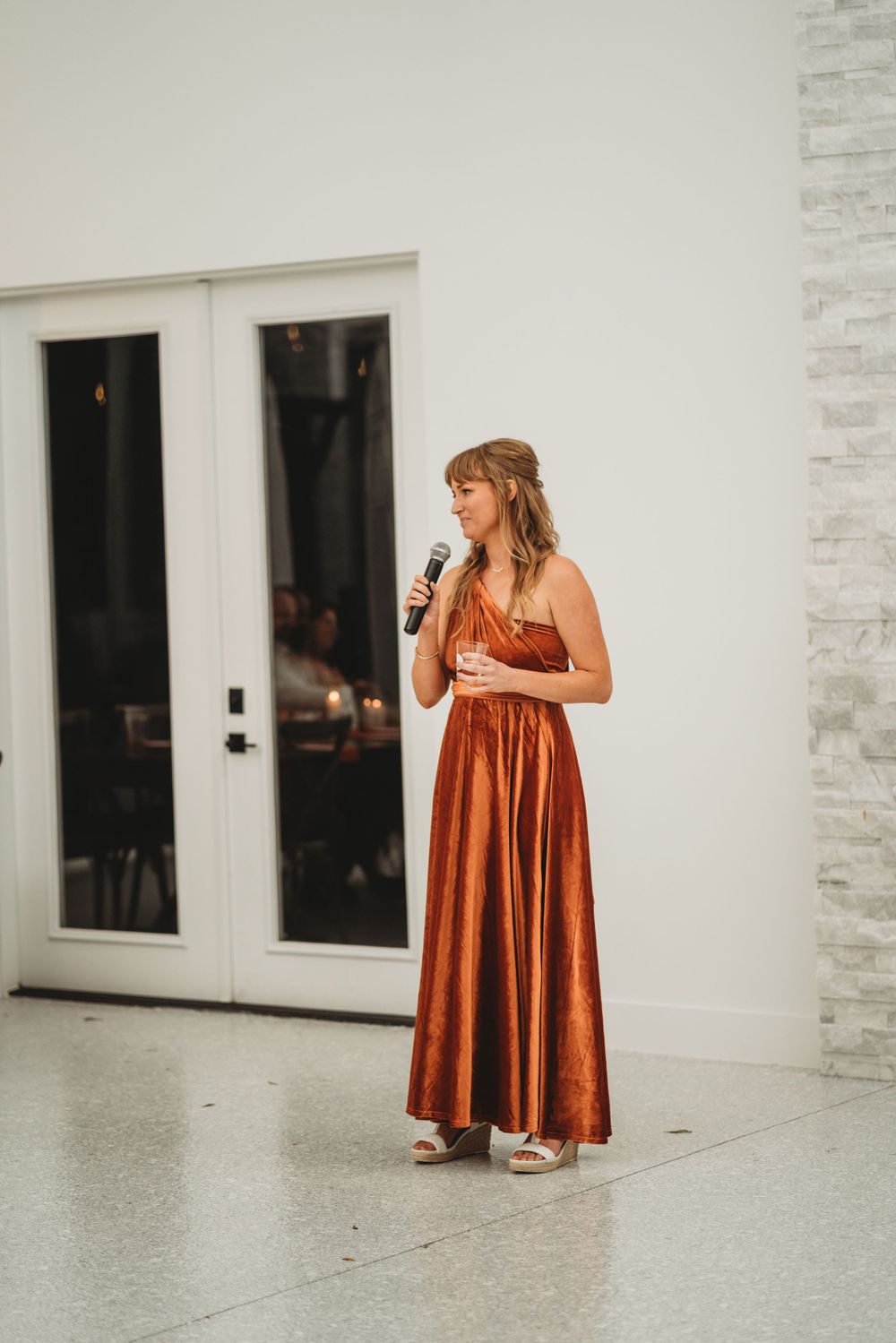 Speaker in rust-colored formal dress giving a toast against white interior walls.