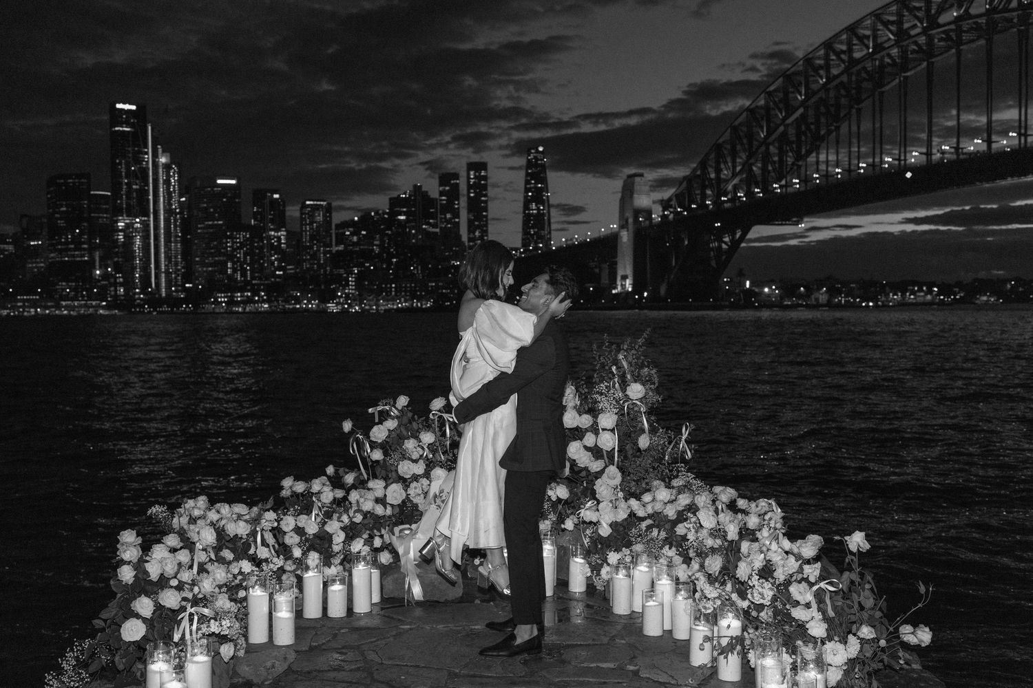 Illuminated paper lanterns line a waterfront path at night with a bridge visible in the background.