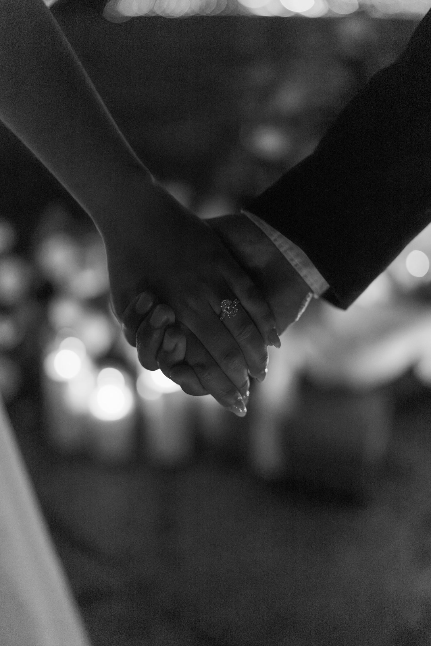 Black and white close-up image of hands holding a ring with bokeh lights in the background.