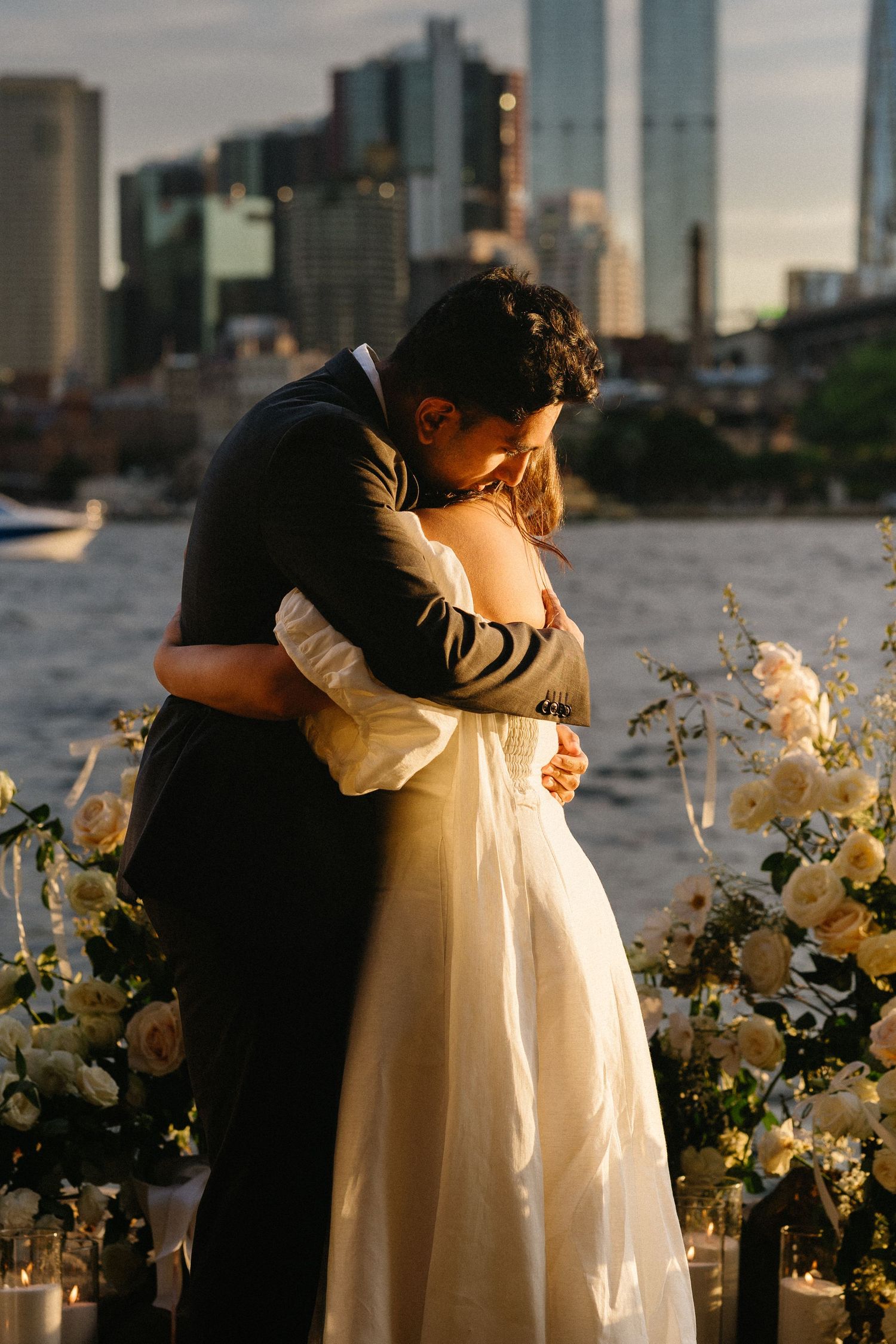 A romantic couple embraces during sunset with white floral arrangements and a city skyline in the background.