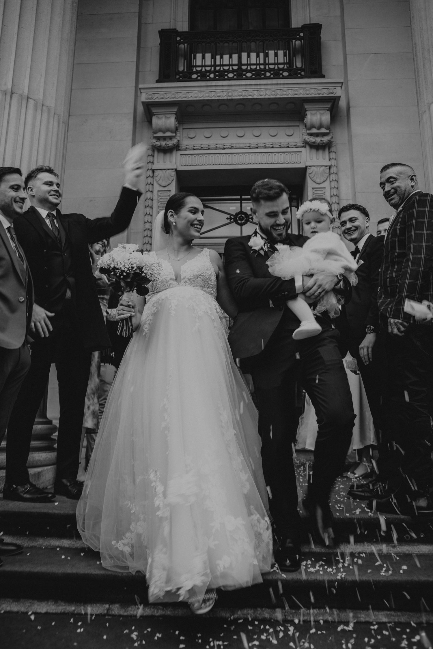 A joyful wedding party celebrates on church steps amid falling confetti.