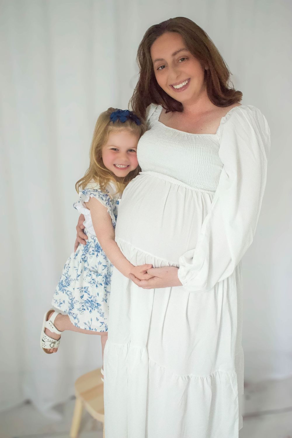 Mother in white dress and daughter in floral dress pose for maternity photo.