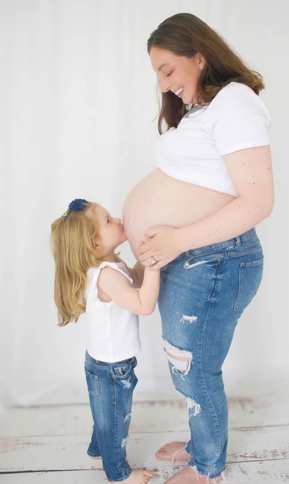A pregnant woman in white shirt and jeans shares a tender moment with a small child.