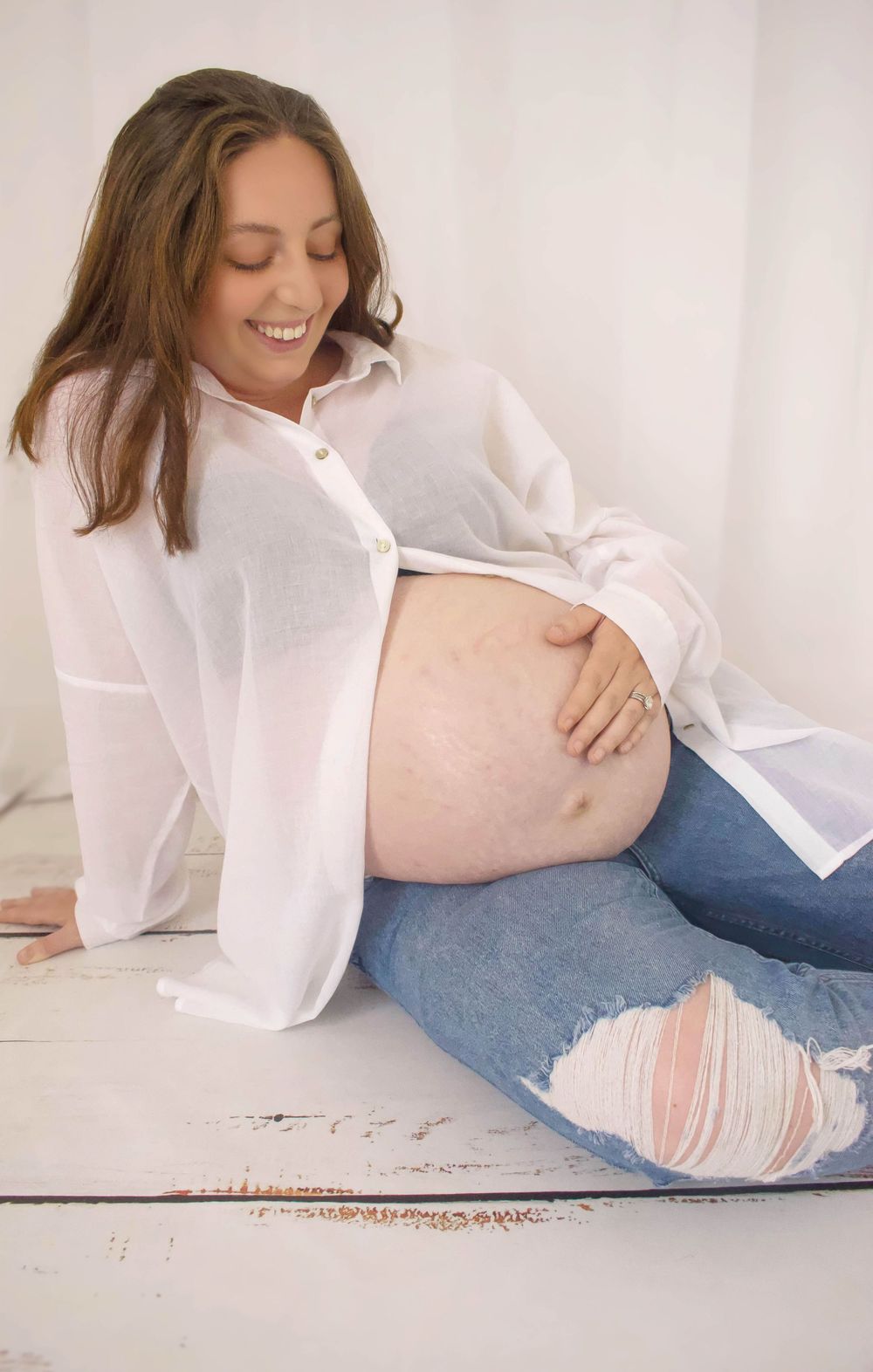 Expectant mother in white shirt and distressed jeans smiles while touching her baby bump.
