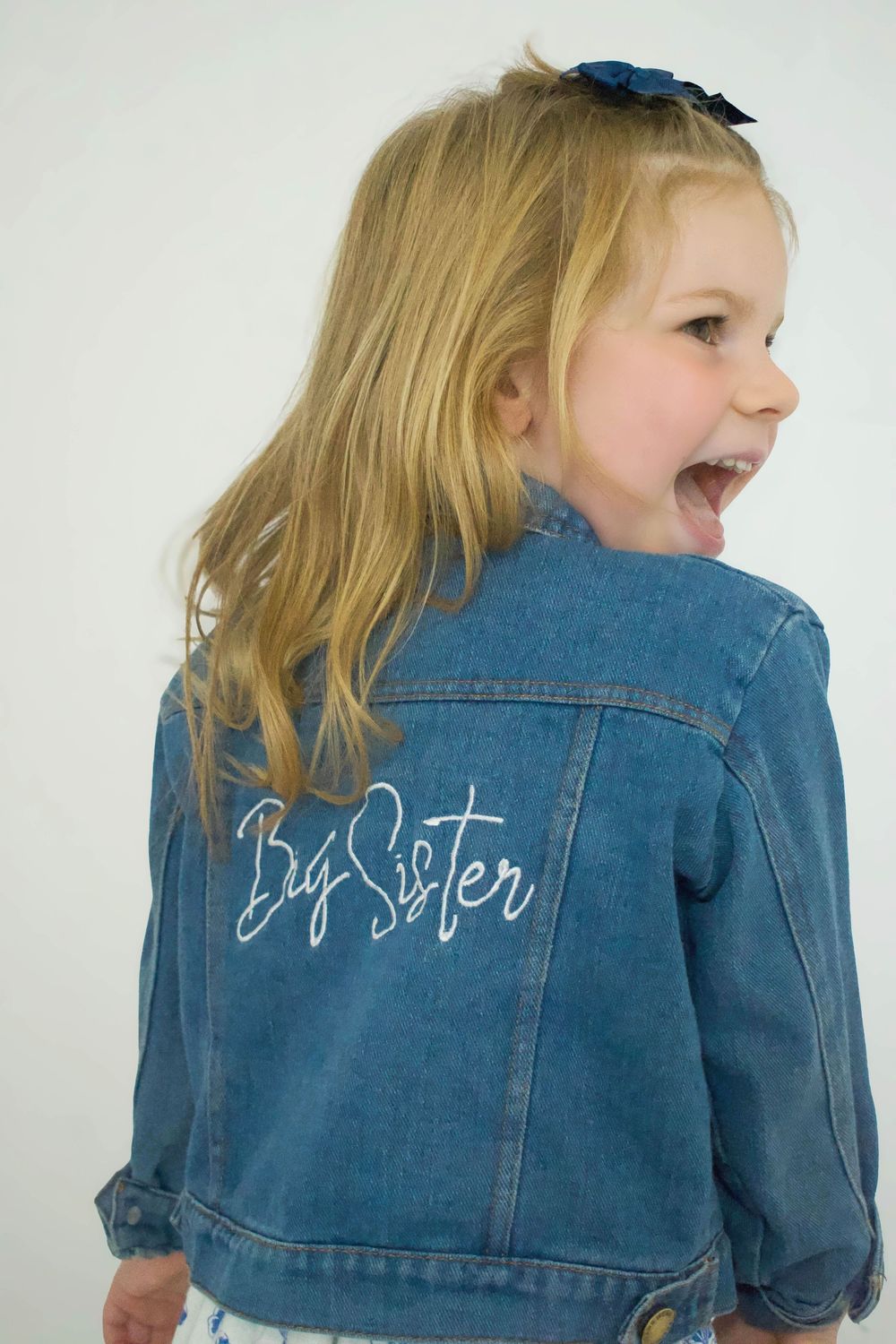 Young child wearing a denim jacket with 'Big Sister' text laughs joyfully.