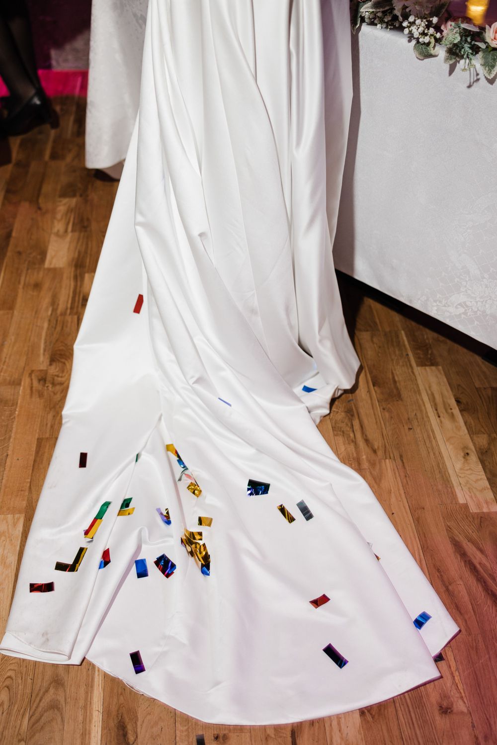 White wedding dress train decorated with colorful confetti on wooden floor.