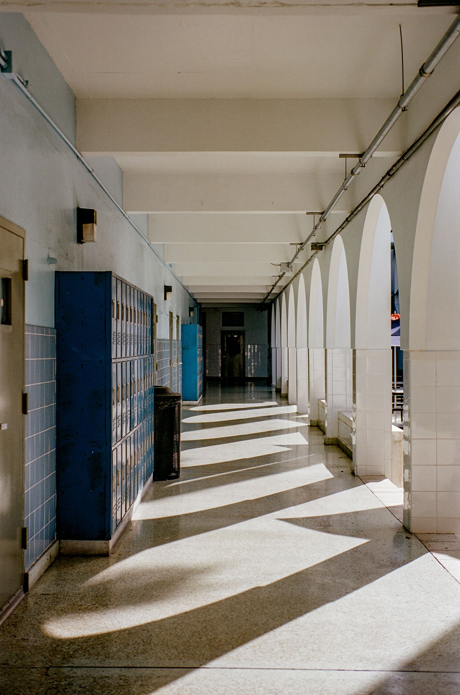 Long institutional hallway with lockers showing dramatic shadows from arched windows.