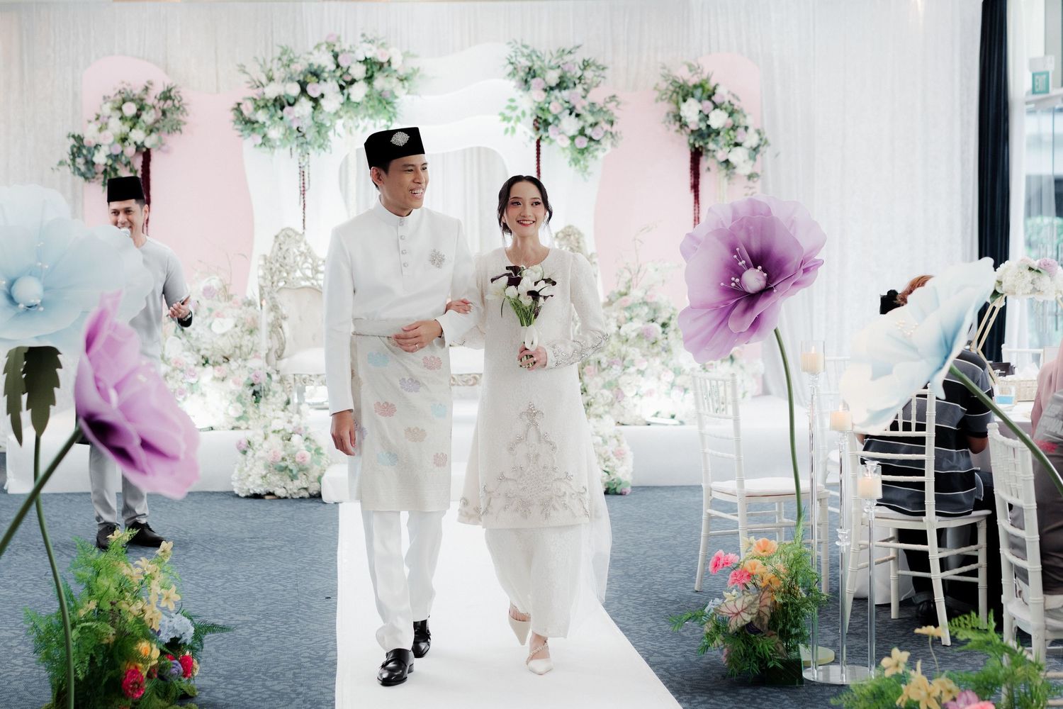A couple walks down the aisle at an elegant white floral wedding ceremony with purple flower decorations.