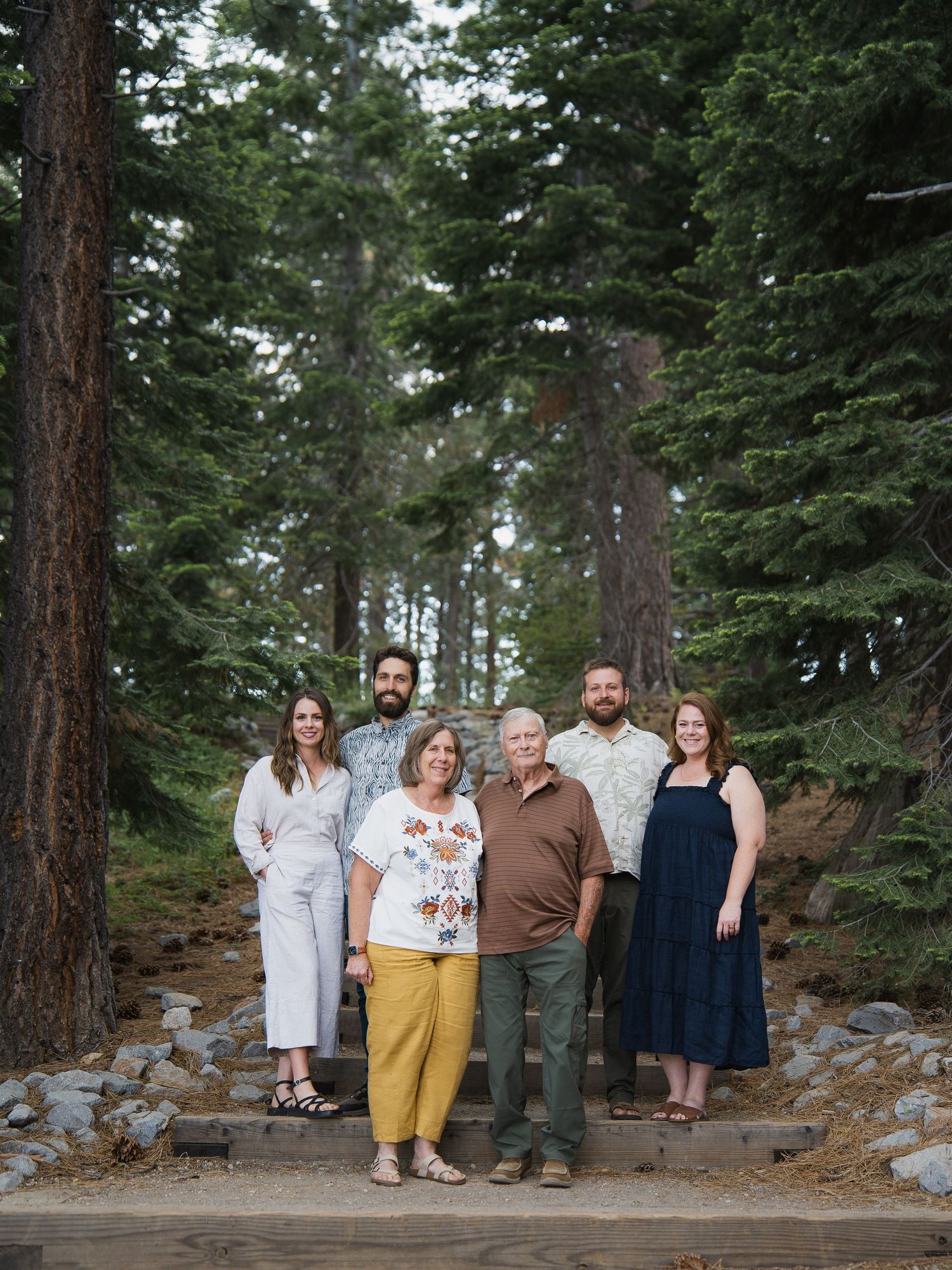Family of six posing outdoors among tall pine trees on stone steps in a forest setting.
