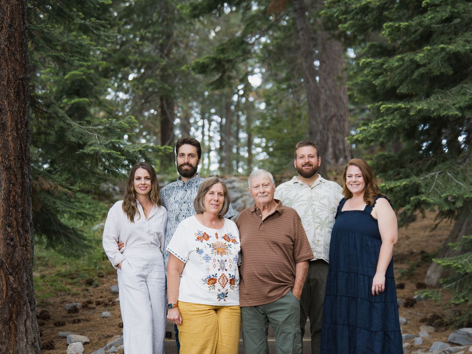 Family of six posing outdoors among tall pine trees in a forest setting for a professional portrait.