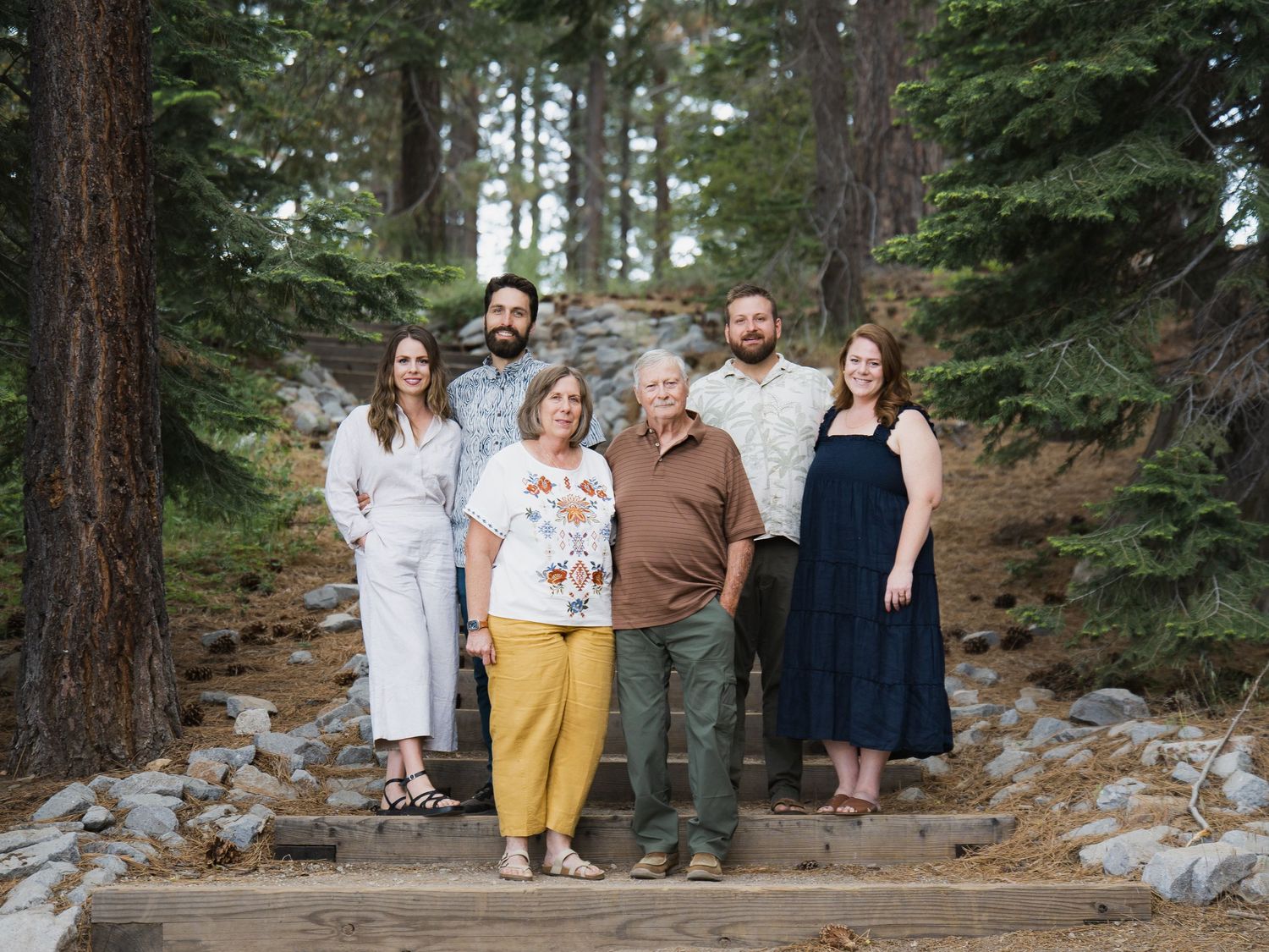 Family of six posing on wooden steps surrounded by tall pine trees in a forest setting.