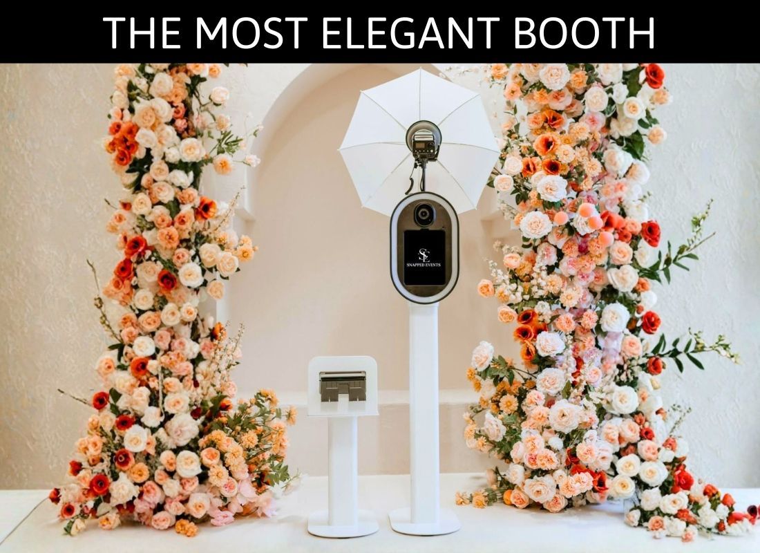 Elegant photo booth stand surrounded by lush floral arrangements of pink and white roses at a wedding venue.