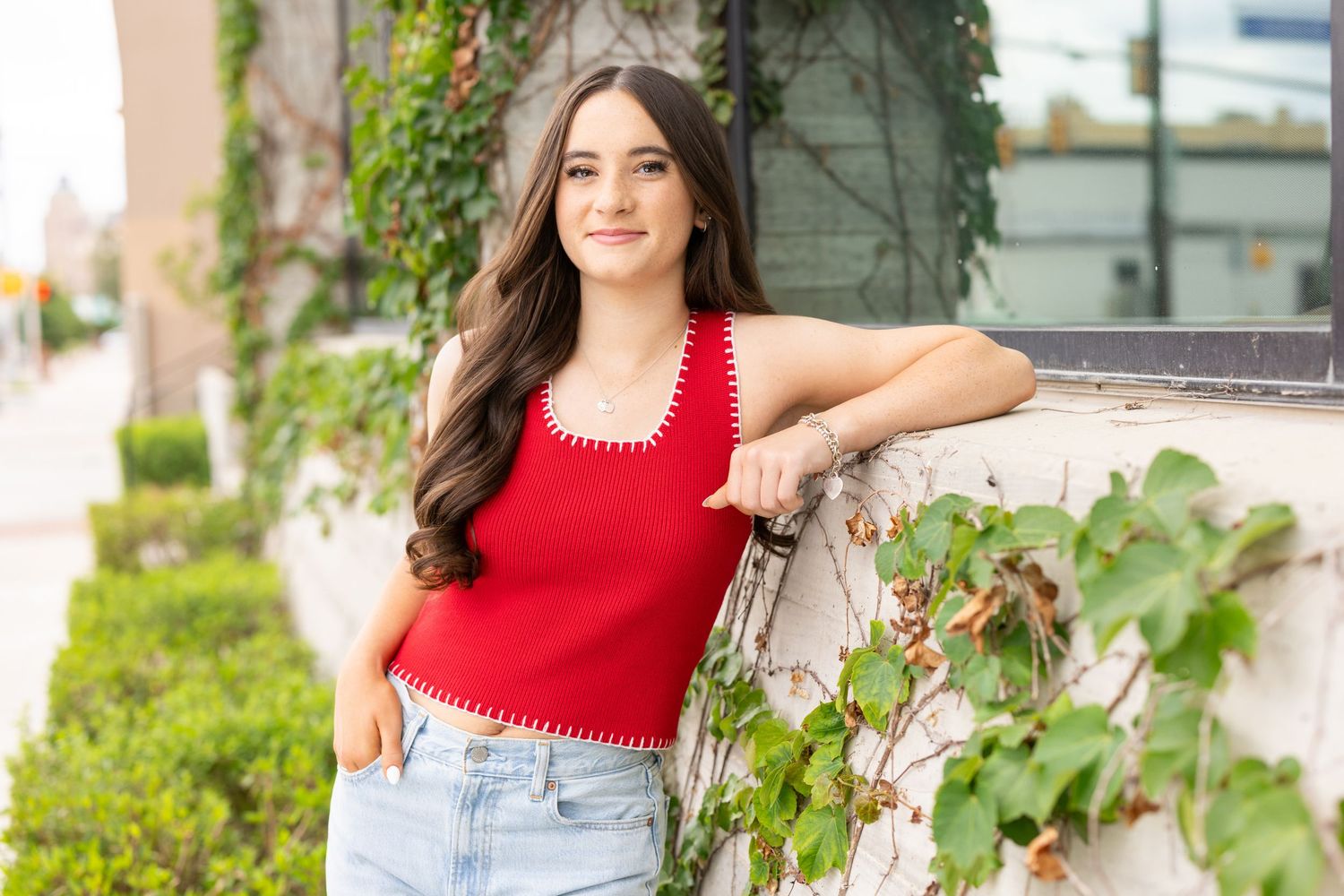 2027 senior girl in red crop top and jeans leaning against a white stone wall with ivy outdoors.