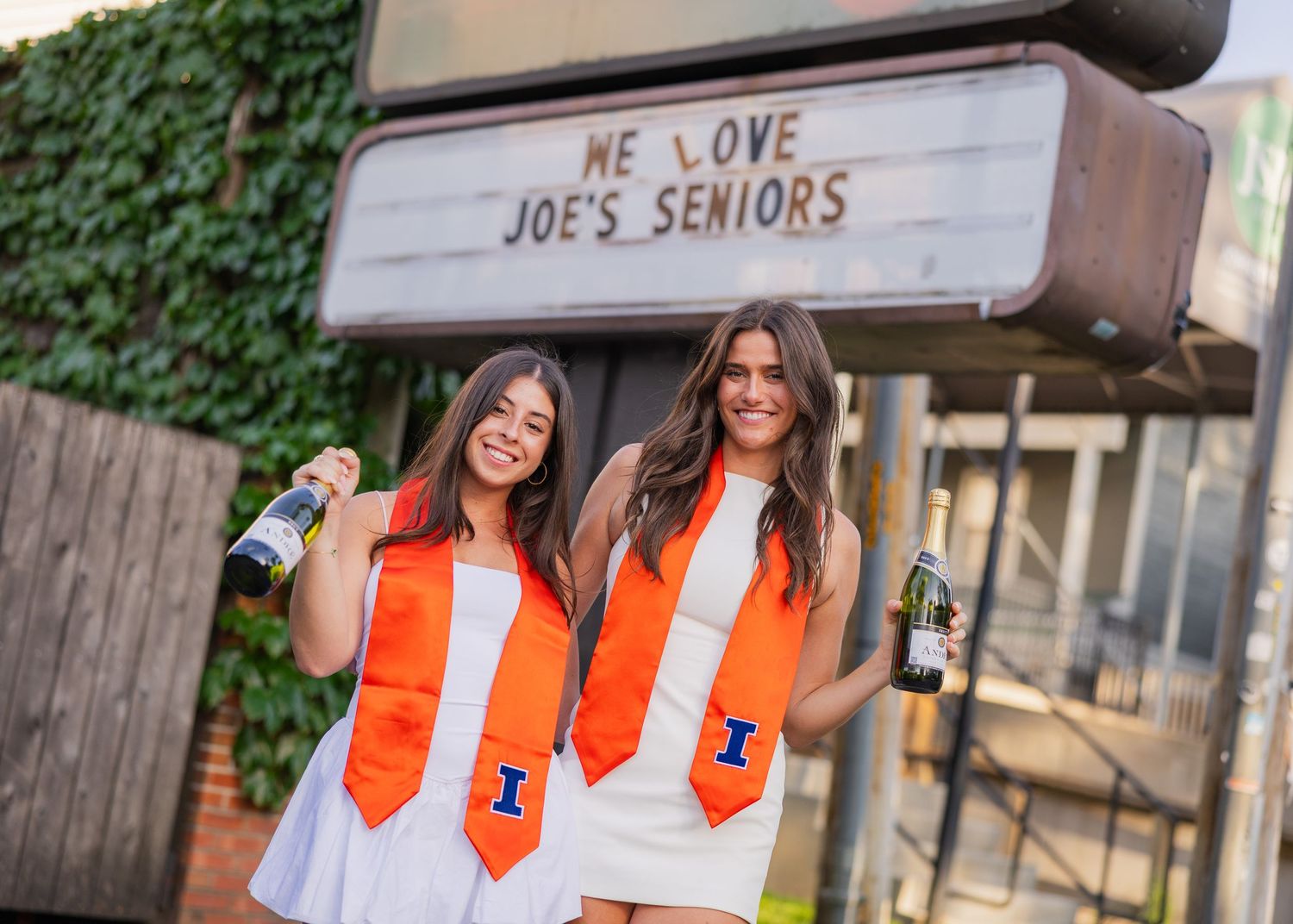 Two graduating girls at Joe's Brewery holding up champagne bottles with University of Illinois stole on