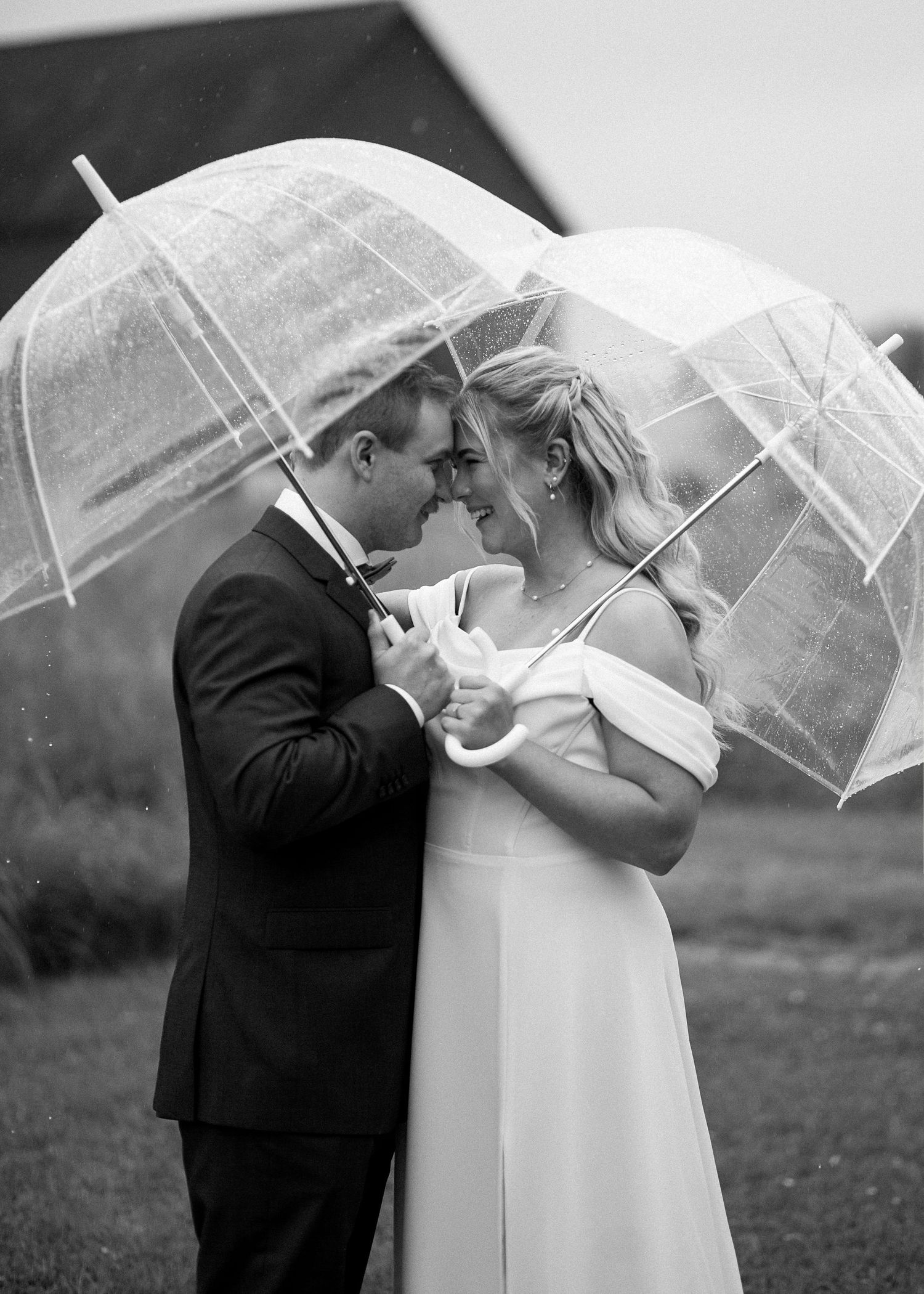 Black and white photo of a bride and groom sharing a romantic moment under clear bubble umbrellas in the rain.