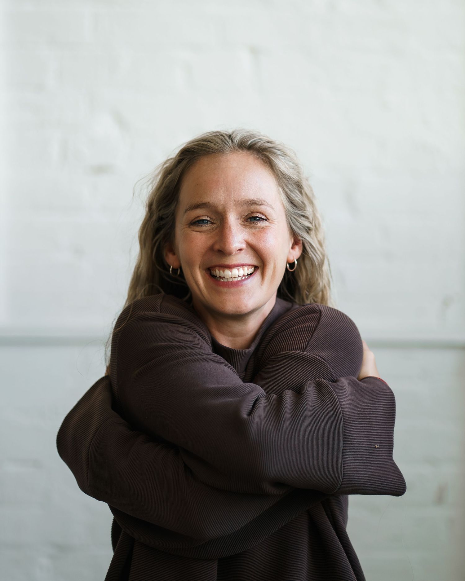 Smiling woman with blonde hair hugging herself, wearing a dark brown hoodie, standing against a white brick wall.
