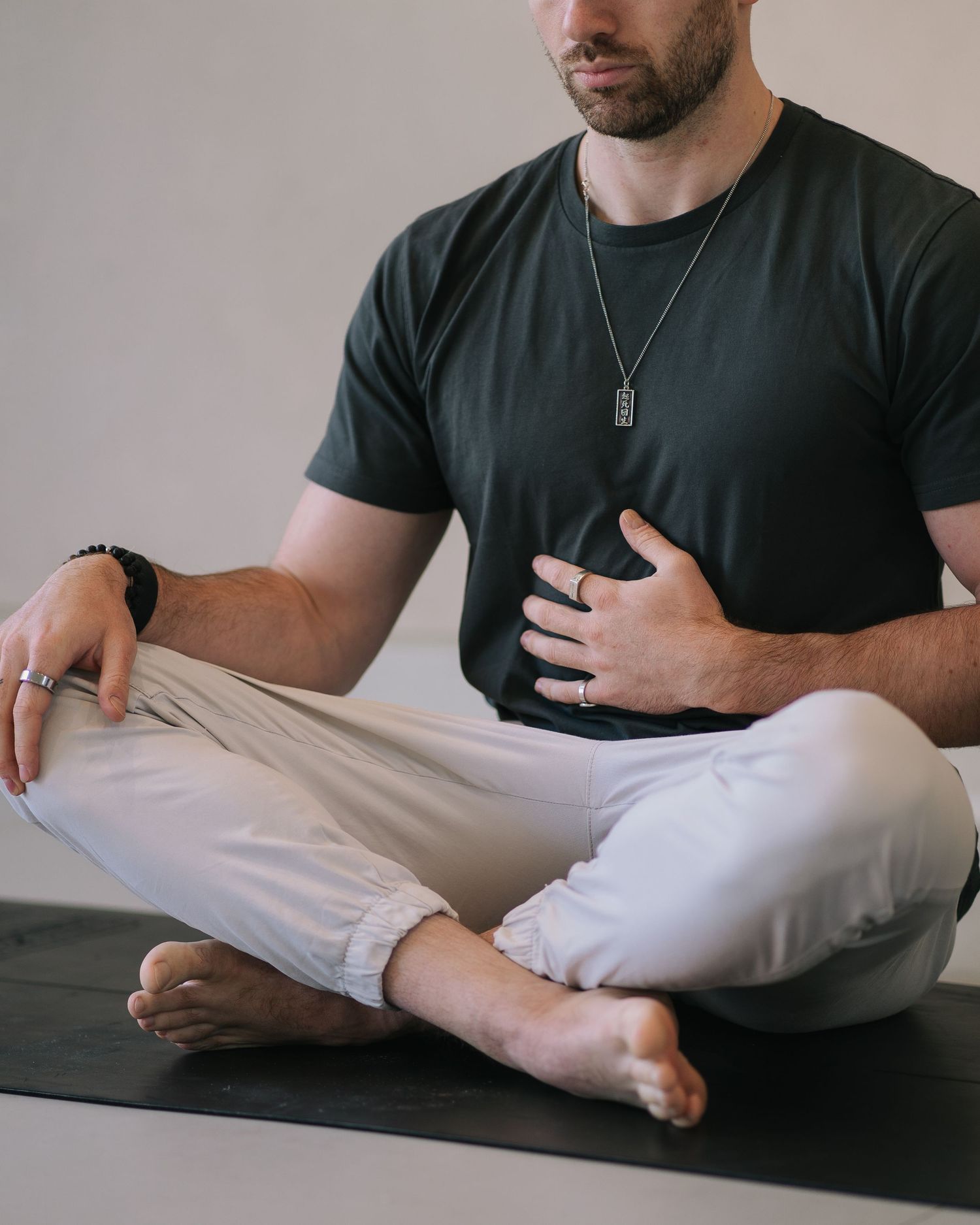 Man in black t-shirt and beige pants sitting cross-legged on yoga mat in meditative pose wearing necklace and bracelet.