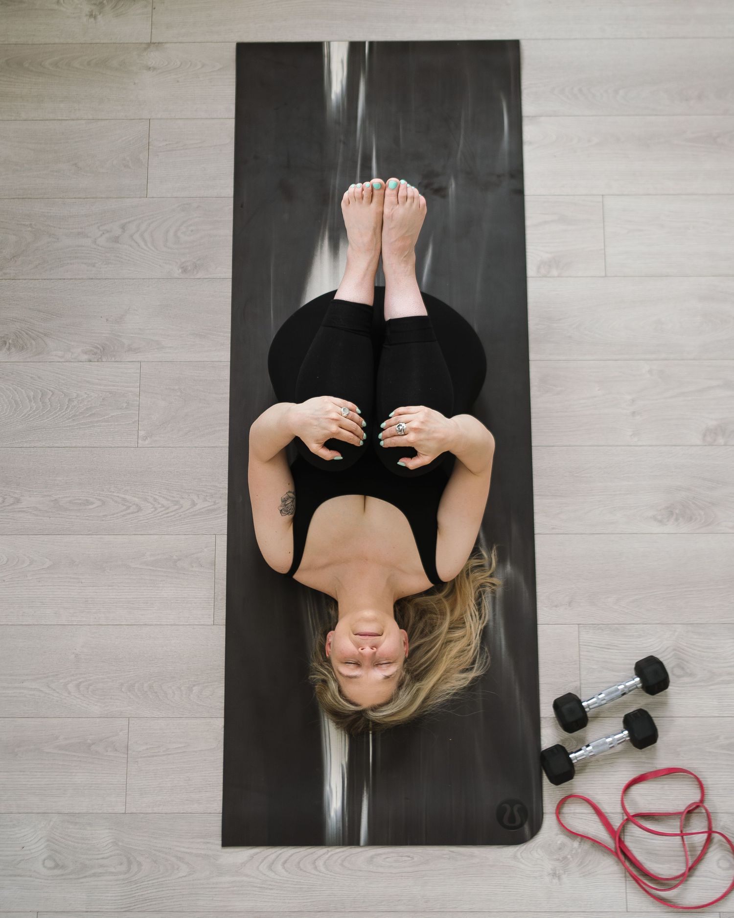 Woman lying on black yoga mat in workout attire with dumbbells and resistance band nearby on wooden floor.
