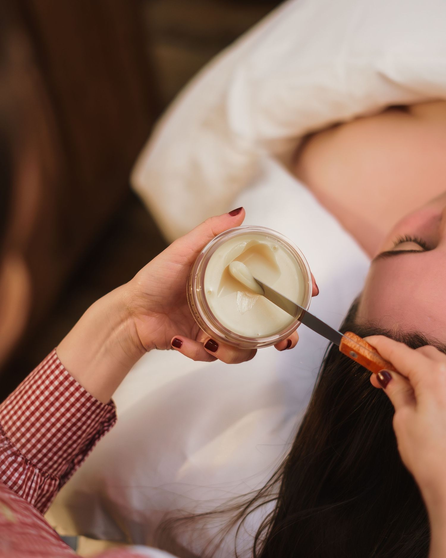 Esthetician applying white cream from a jar with a spatula to a reclining client's face during a facial treatment.