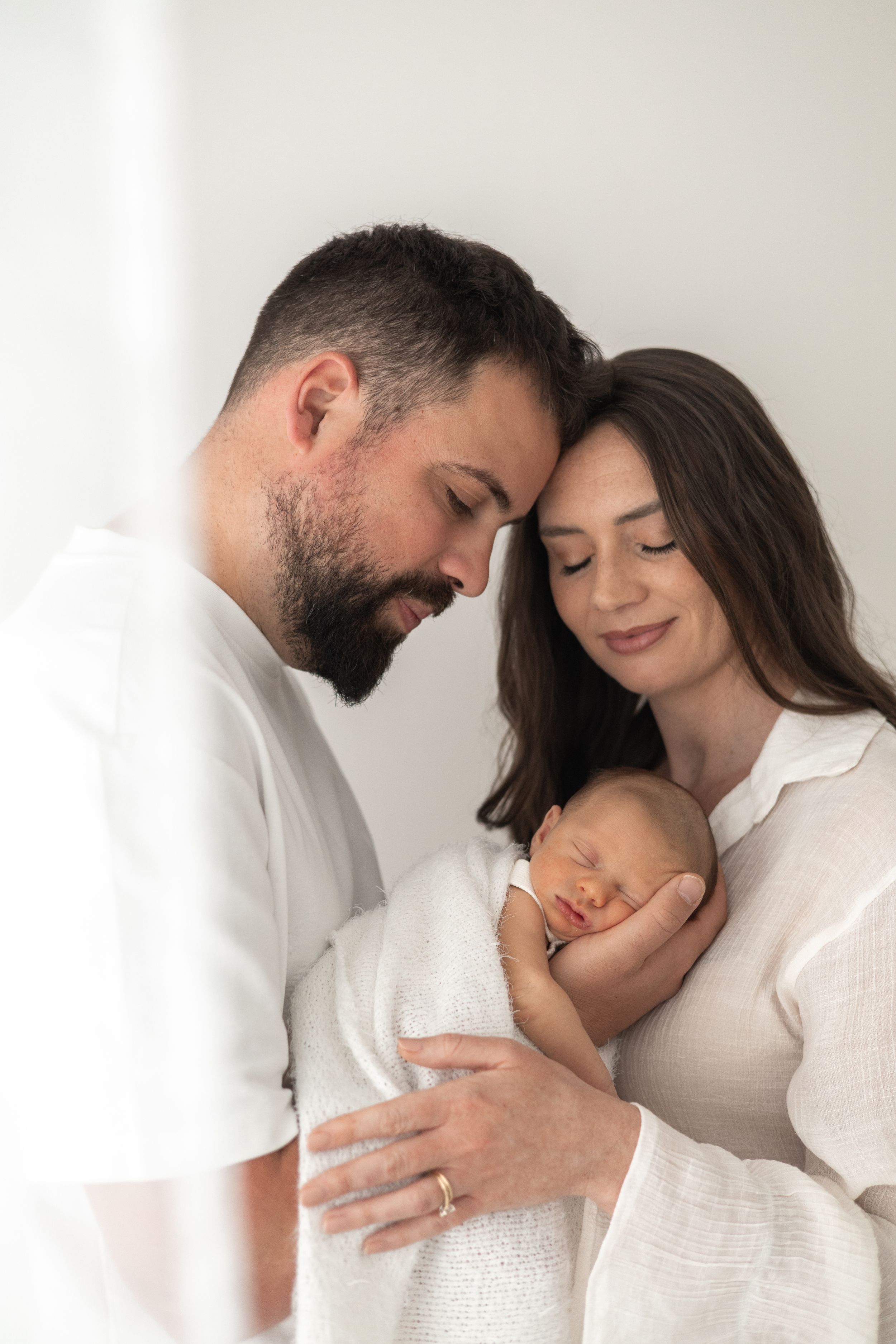 Mother and Father tenderly cradles their sleeping newborn baby against a soft white background Nottingham newborn photographer
