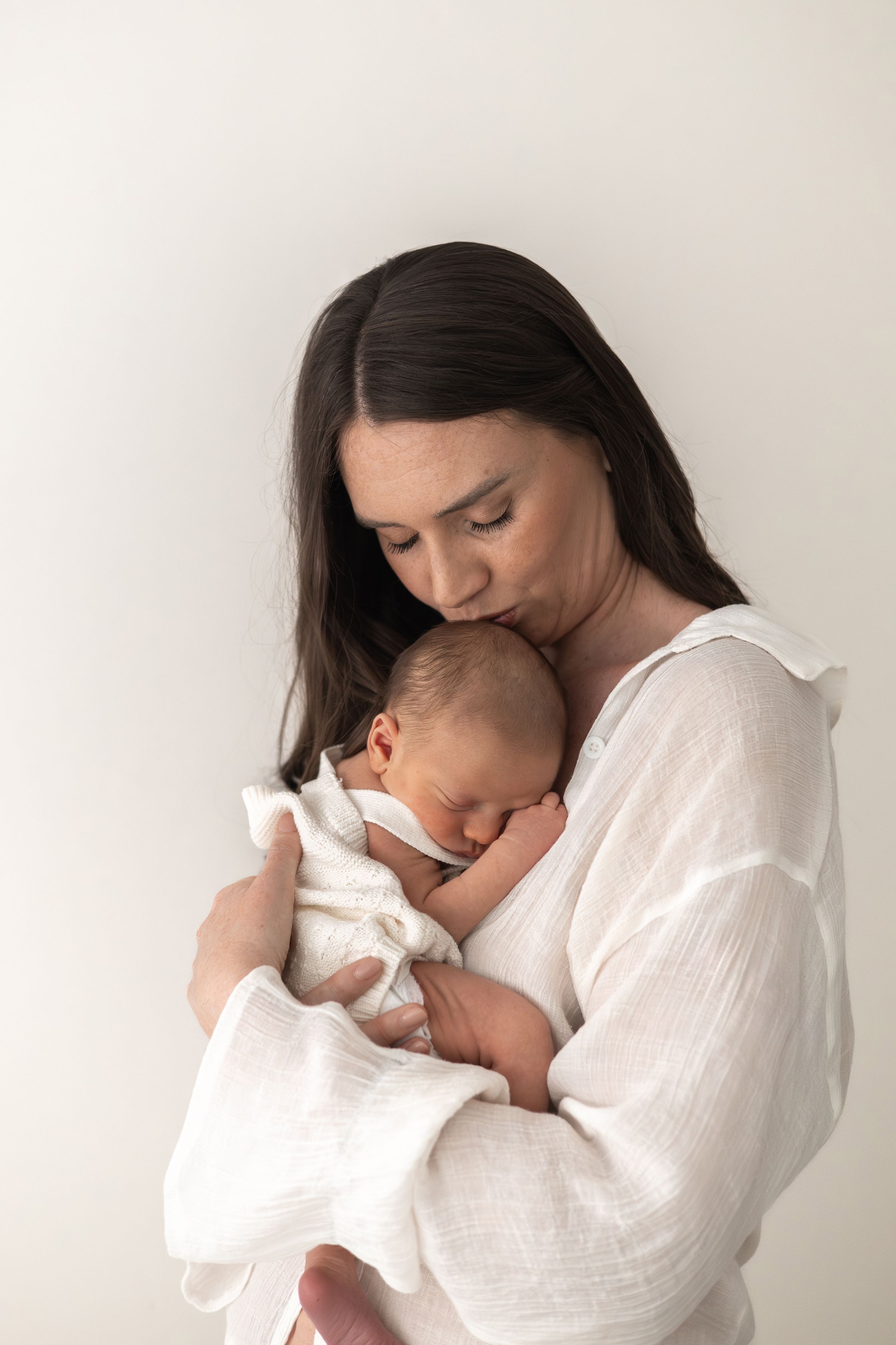 A mother in white linen gently kisses her sleeping newborn baby wrapped in a soft white blanket Nottingham newborn photographer