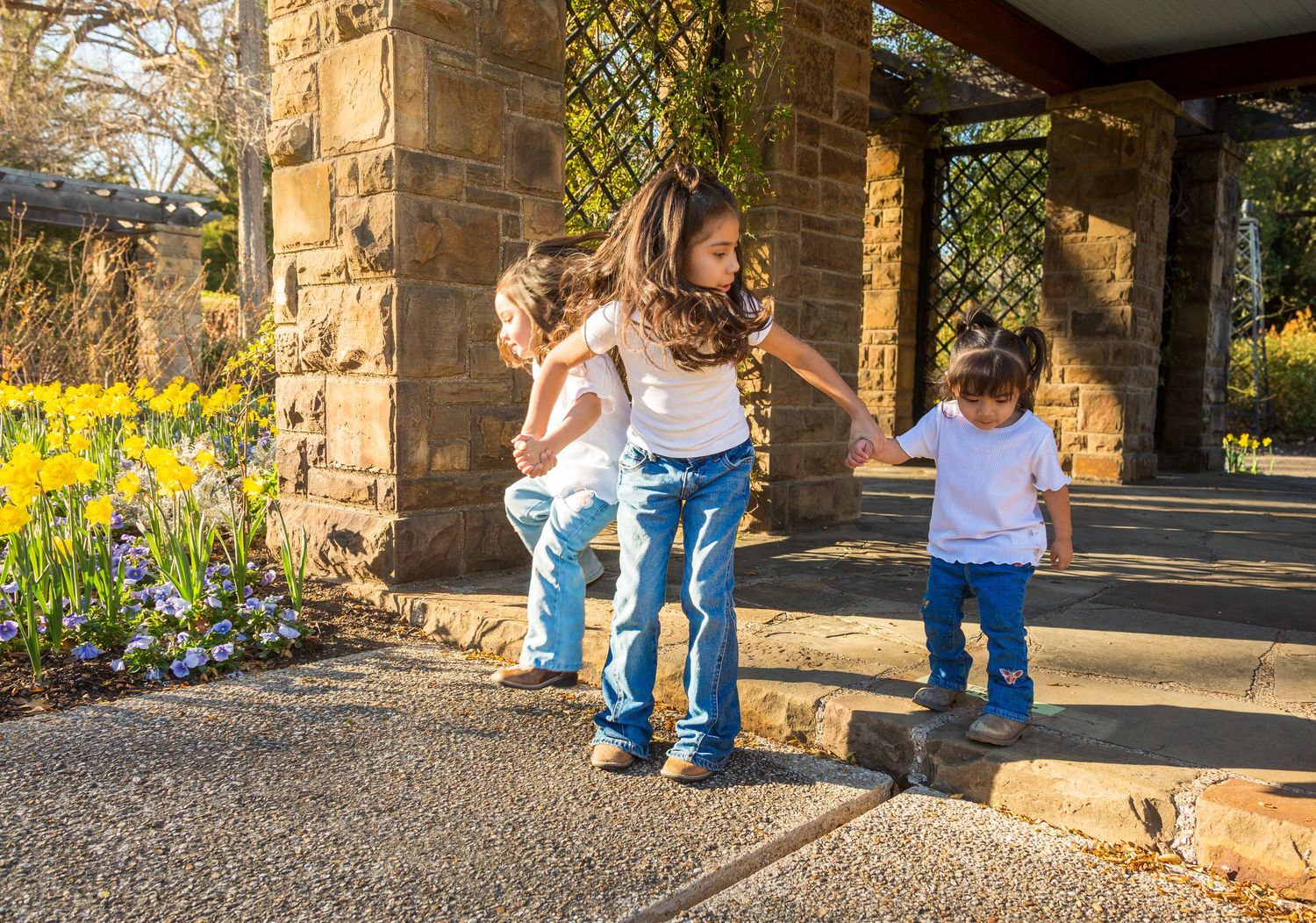 Three children playing outdoors near stone pillars surrounded by blooming daffodils on a sunny spring day.