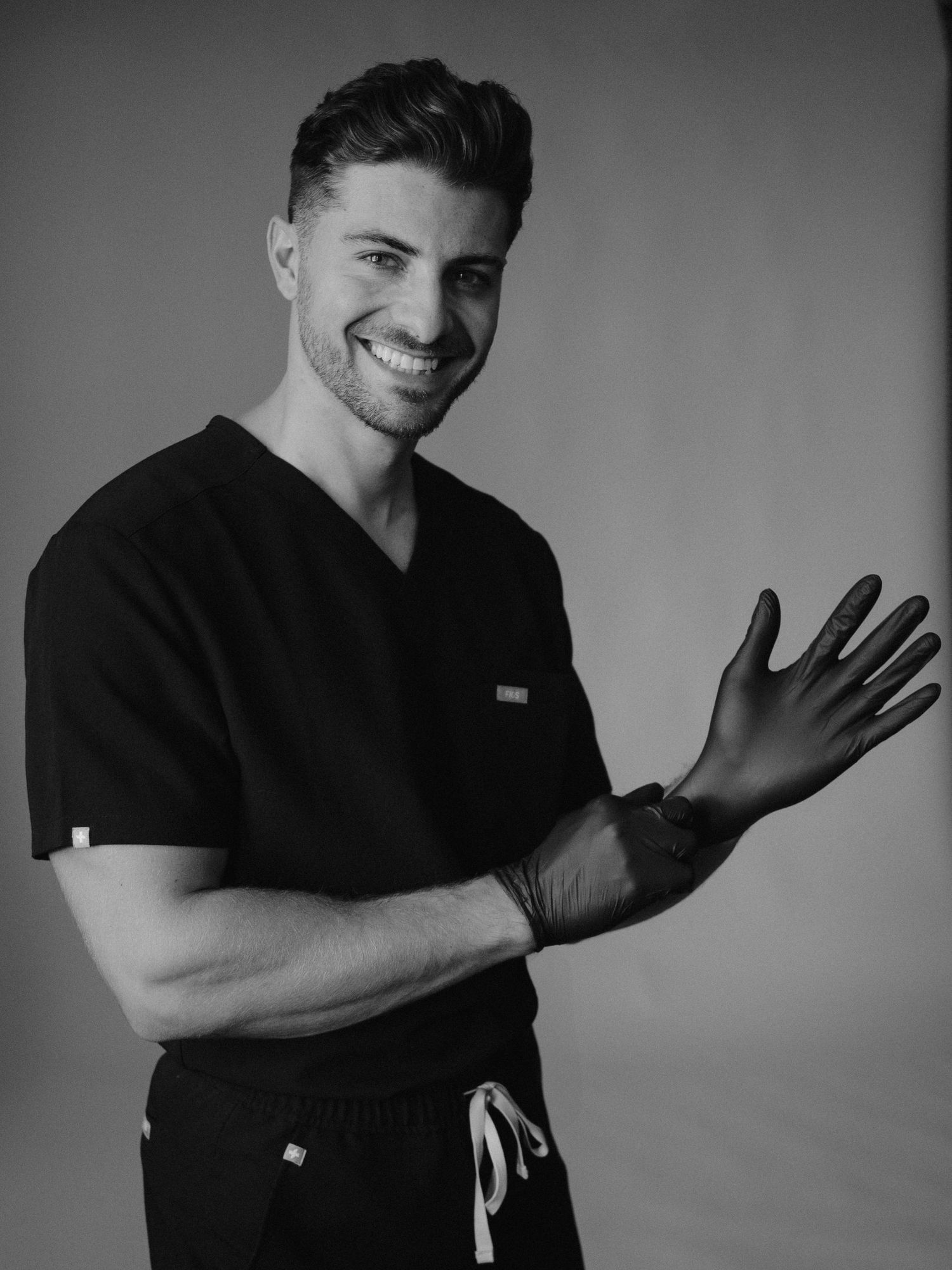 Smiling male healthcare professional in black scrubs putting on a glove, black and white portrait photo.