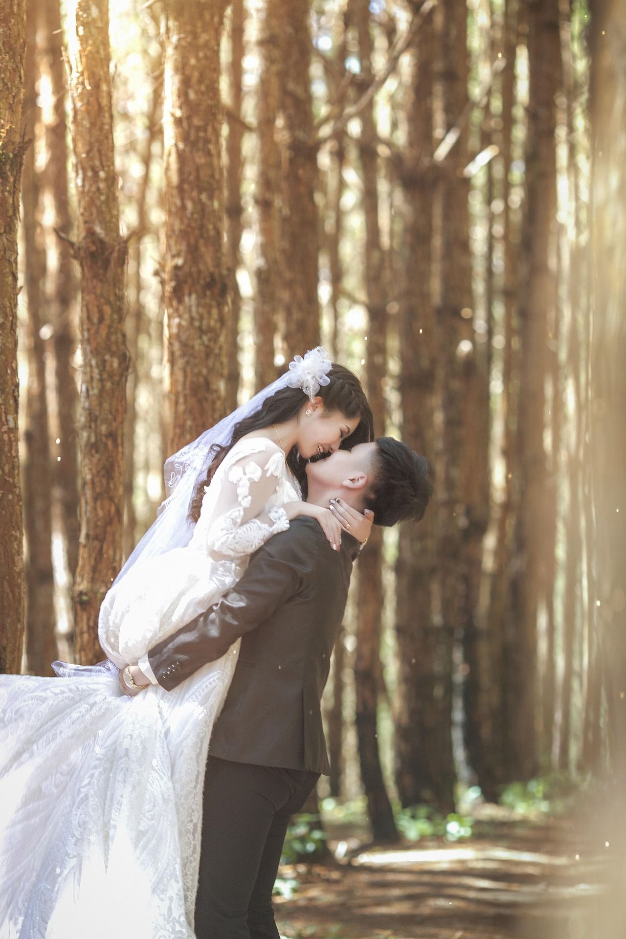 Un marié soulève sa femme pour un baiser romantique au milieu de grands arbres, dans une photo de mariage prise en forêt, baignée de soleil