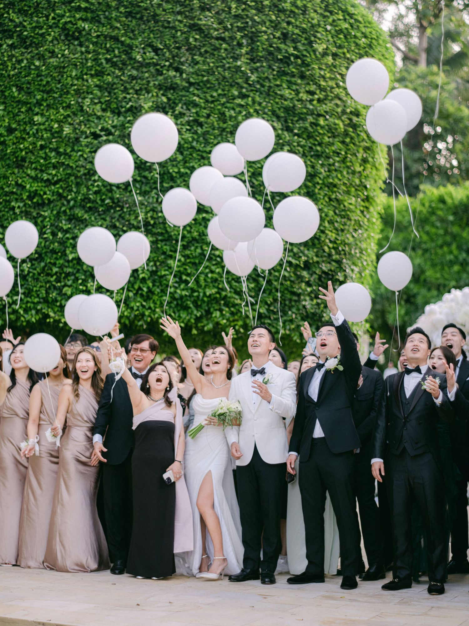 Wedding party releasing white balloons outdoors, bride in white gown with bridal party in blush and black formal attire.