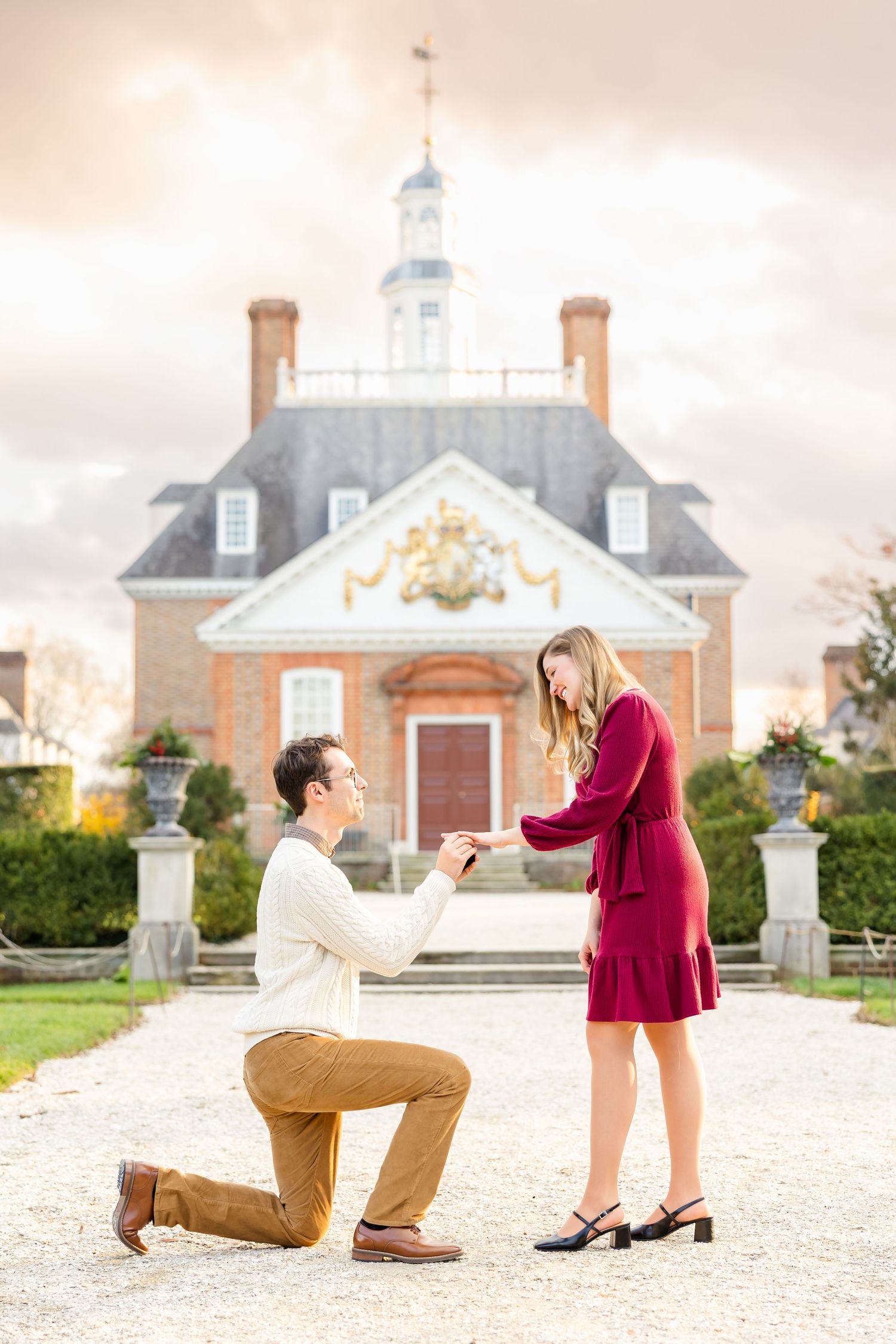 Photograph of a man on one knee proposing to his girlfriend and putting a ring on in front of Governor's Palace in Colonial Williamsburg.