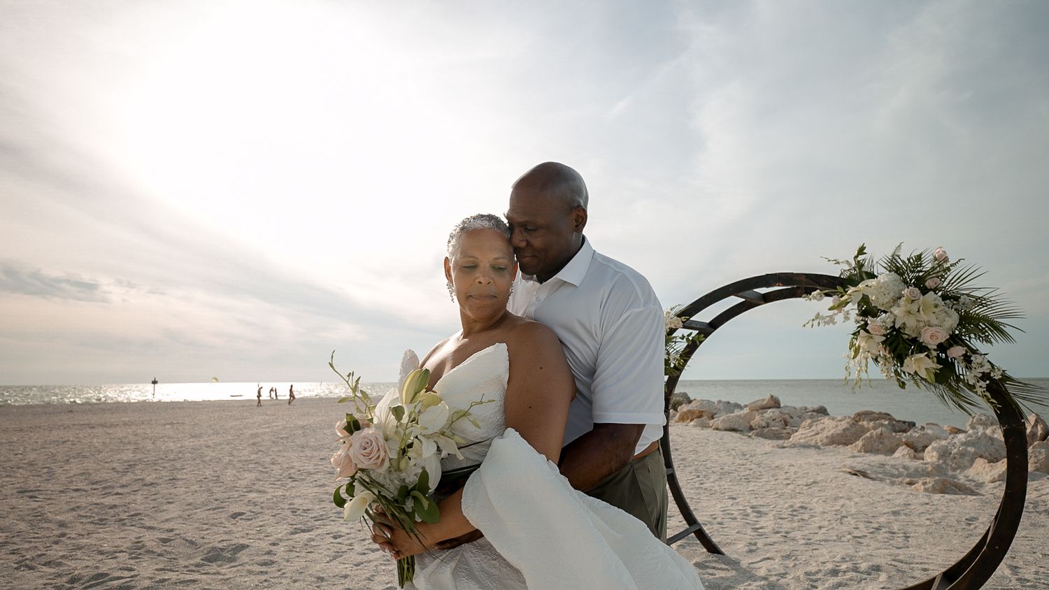 A couple embraces at a beach wedding ceremony with a floral arch, the groom in white and bride holding a bouquet.