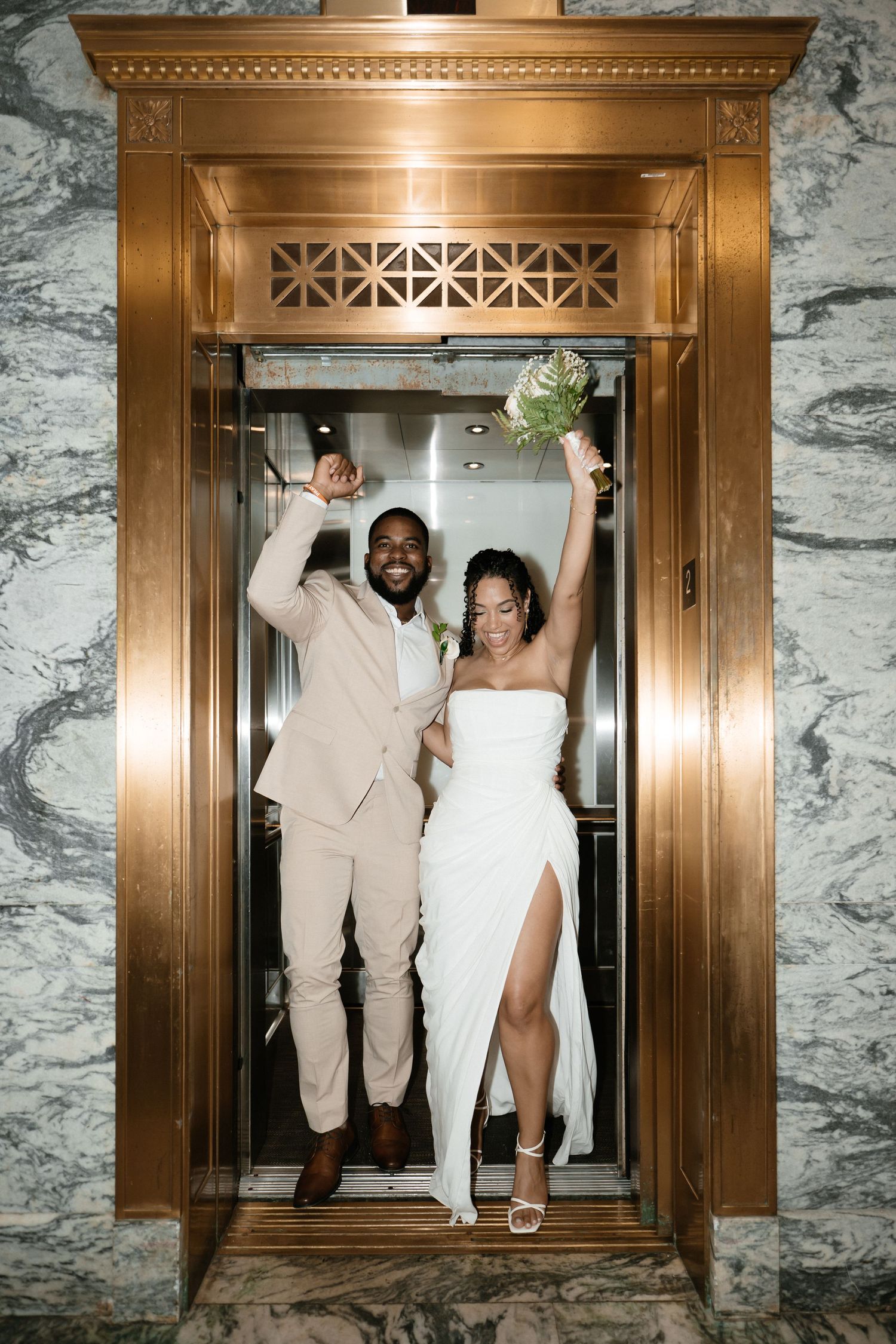 Joyful newlyweds celebrate in a gold elevator, bride in white gown holding bouquet, groom in tan suit, marble walls.