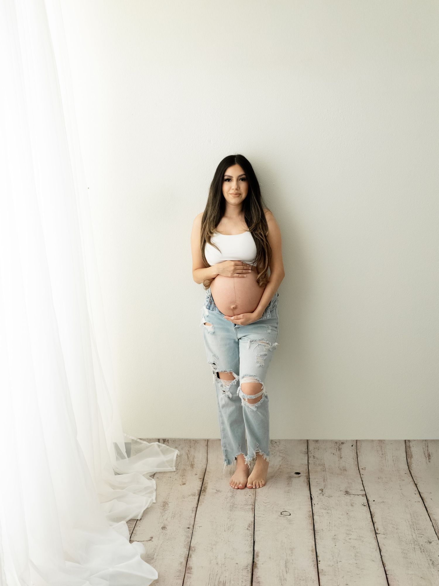 Pregnant woman in white crop top and ripped jeans cradling baby bump against white wall in studio maternity photo.