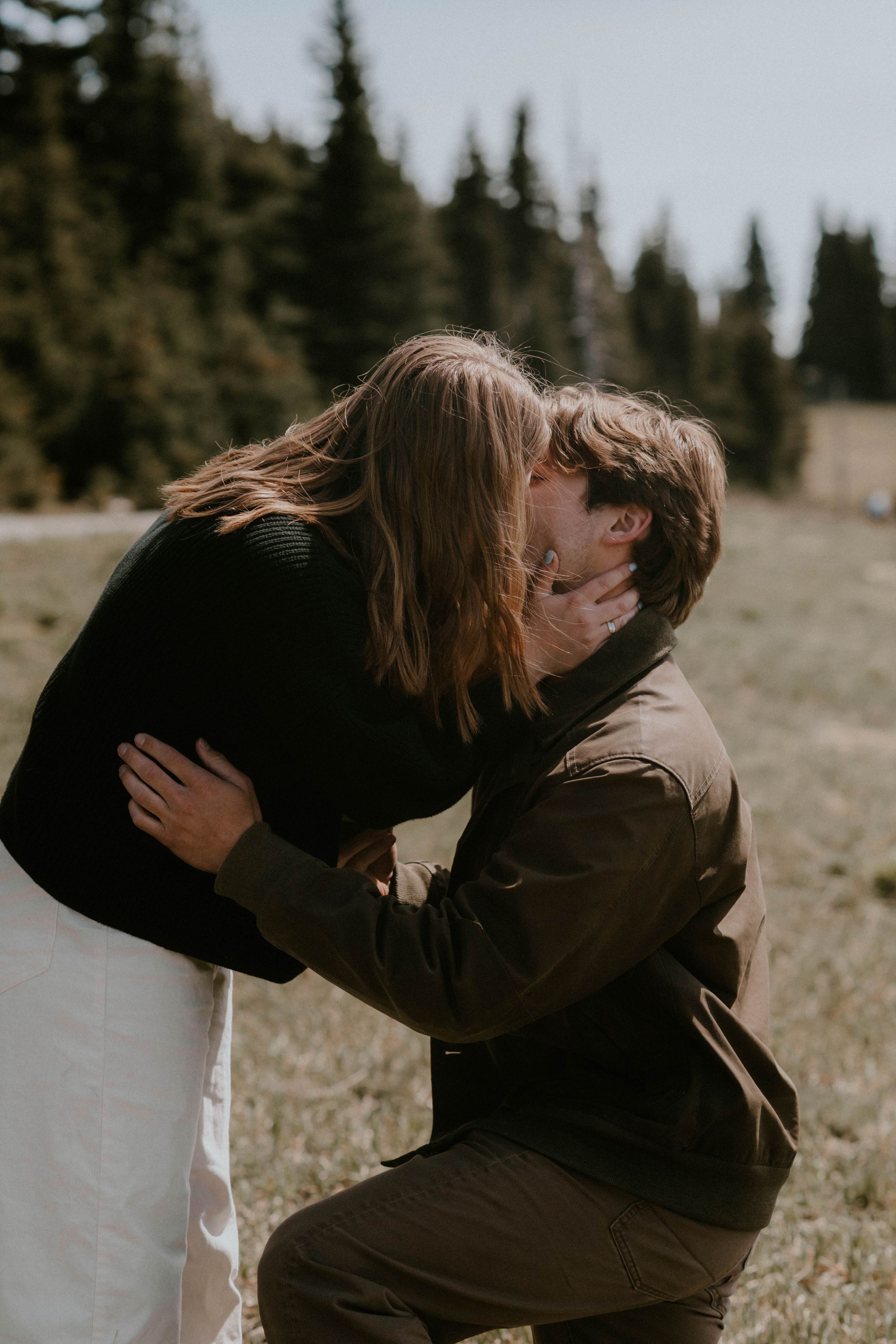 Couple sharing a romantic kiss outdoors in a mountain meadow with evergreen trees in the background.