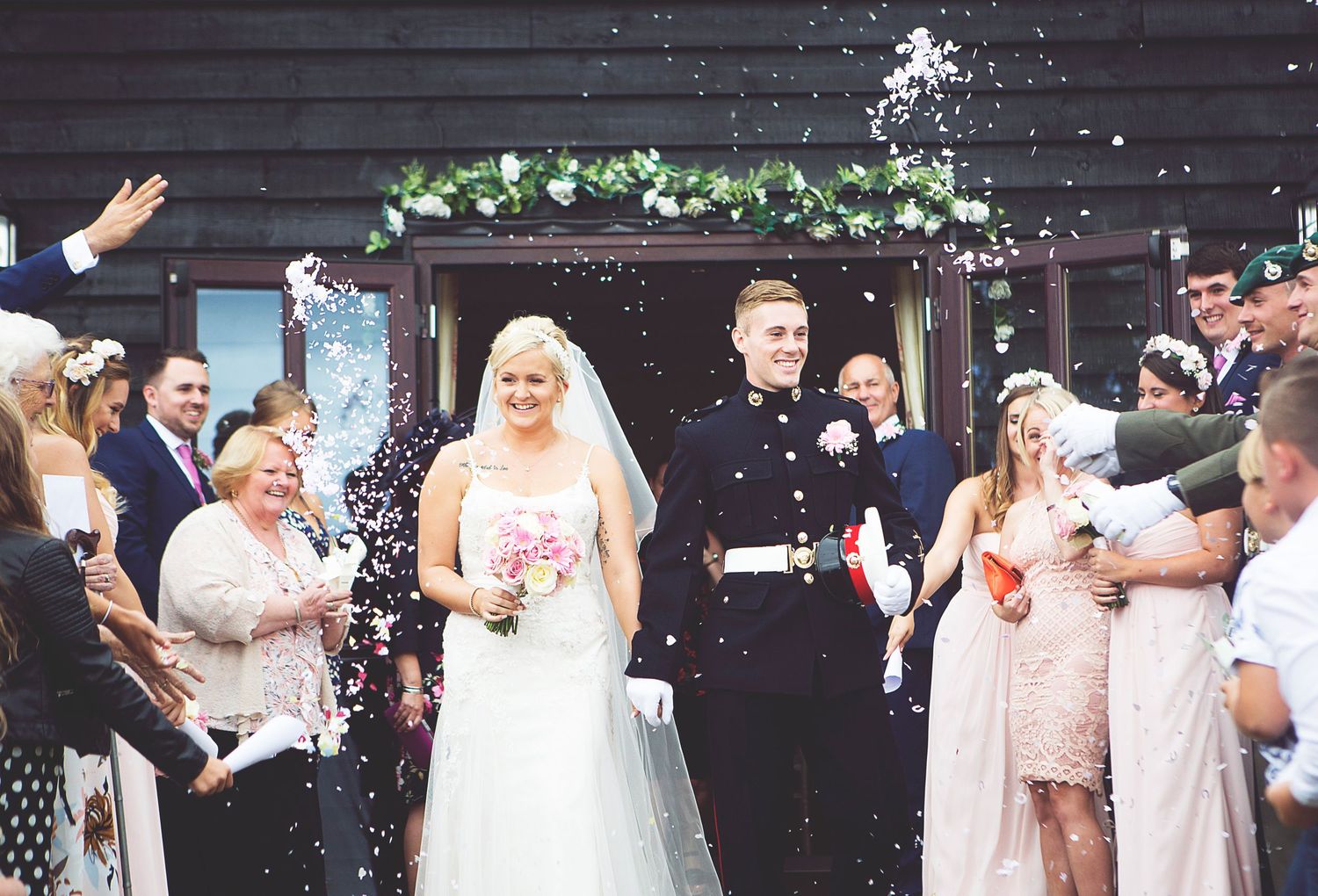 A groom in military uniform kisses the cheek of a smiling blonde bride in a white dress outdoors.