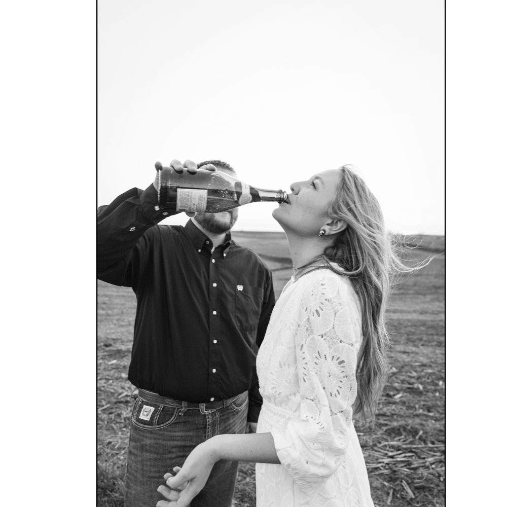 Black and white photo of a couple sharing a drink from a bottle outdoors in an open farm field.