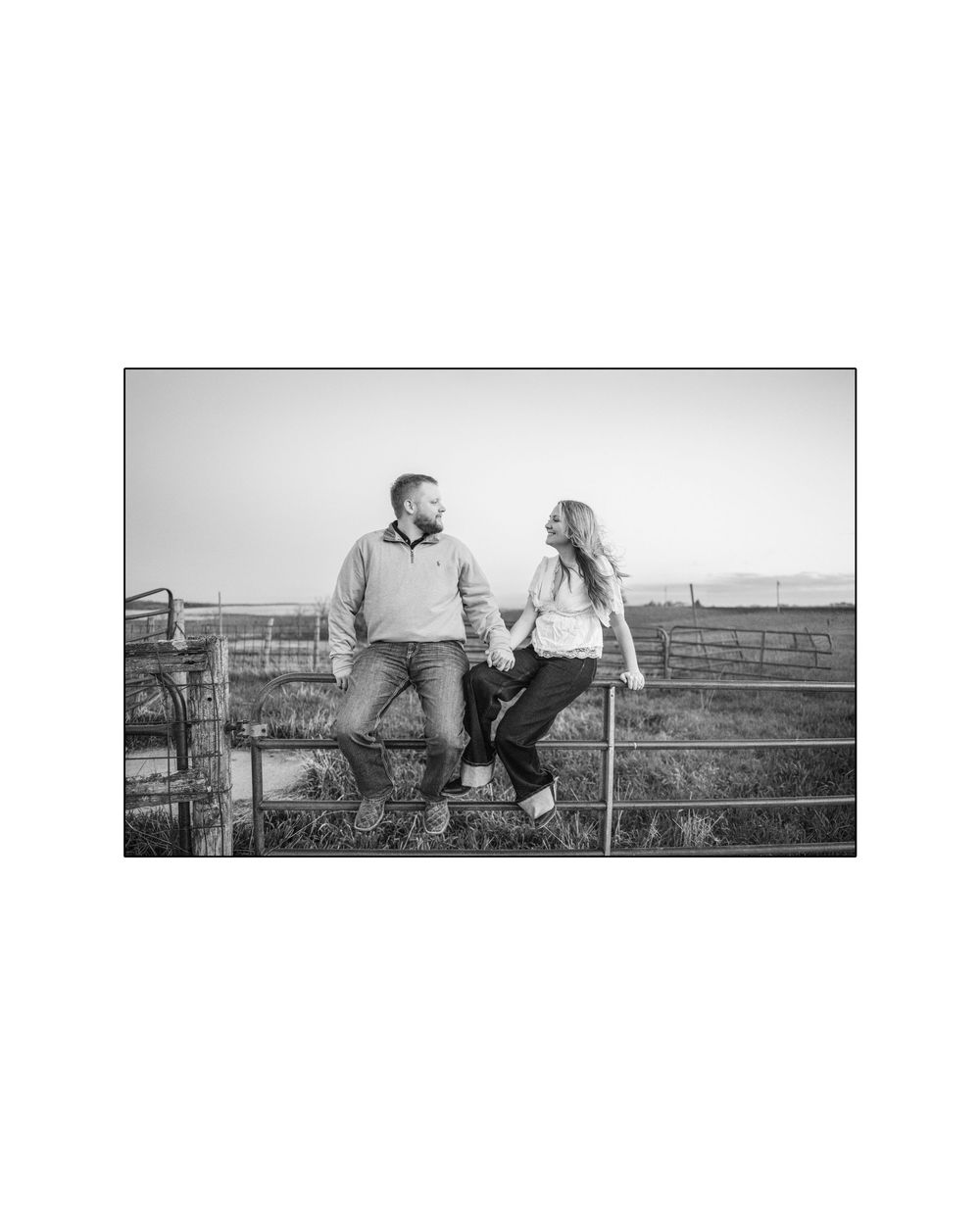 Black and white photo of a couple sitting on a farm gate holding hands, smiling at each other in a rural setting.