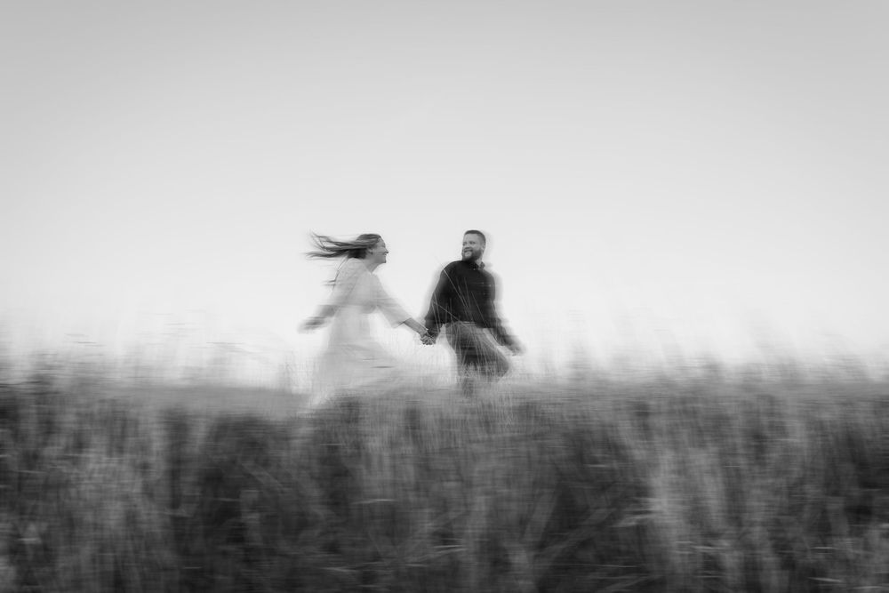 Blurry black and white photo of a couple holding hands running through a field, hair blowing in the wind.