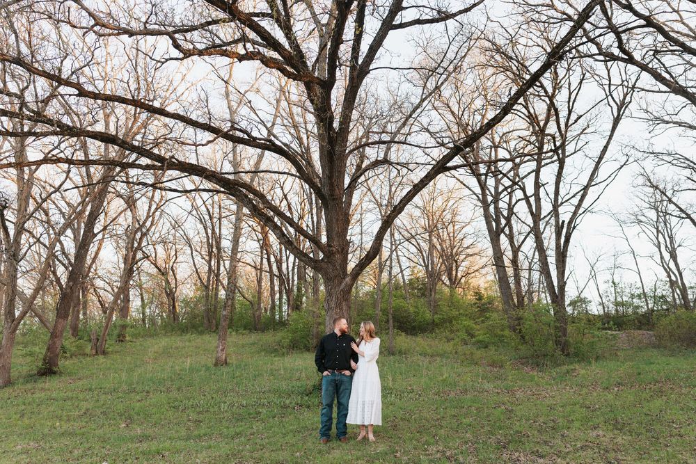 Couple standing together in a lush green meadow beneath a large bare tree at golden hour.