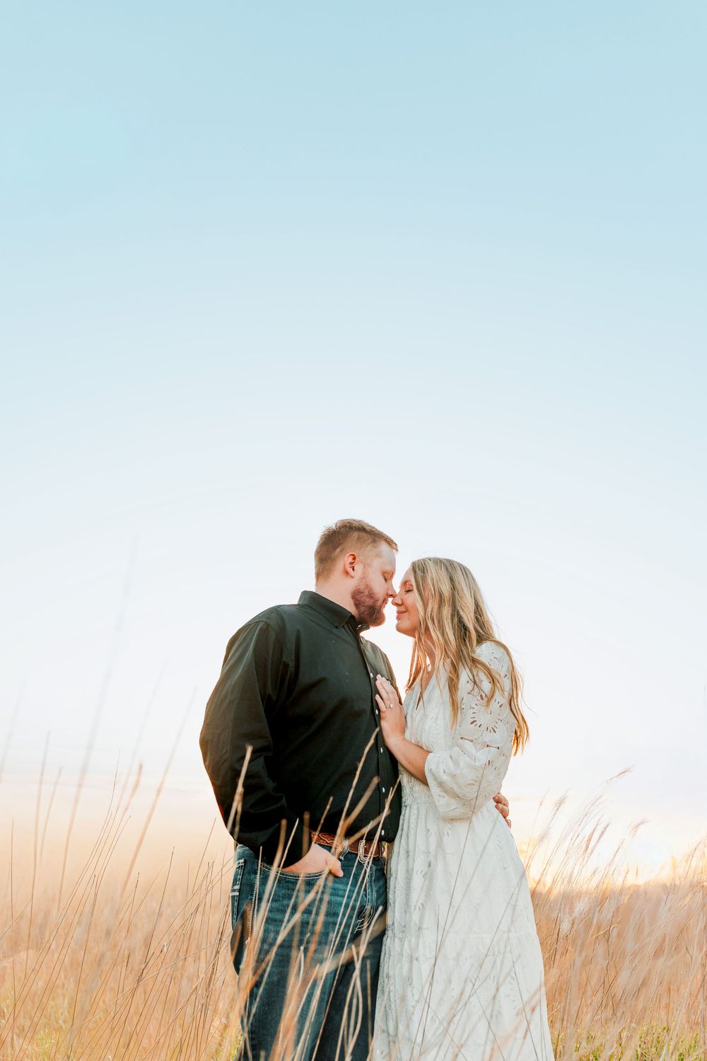 Couple sharing a romantic moment in golden field at sunset, woman in white lace dress and man in black jacket.