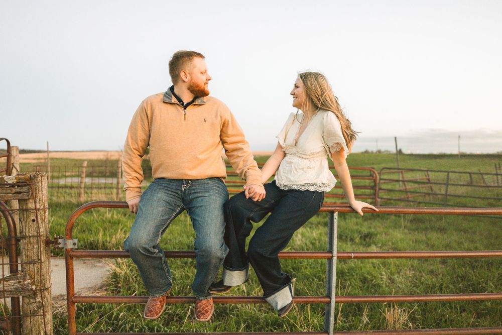 Couple holding hands and smiling at each other while leaning on a rustic red farm gate in a green field.