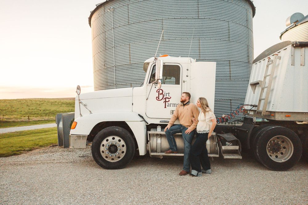 Couple posing by a white farm truck near grain silos at sunset on a rural farm property.