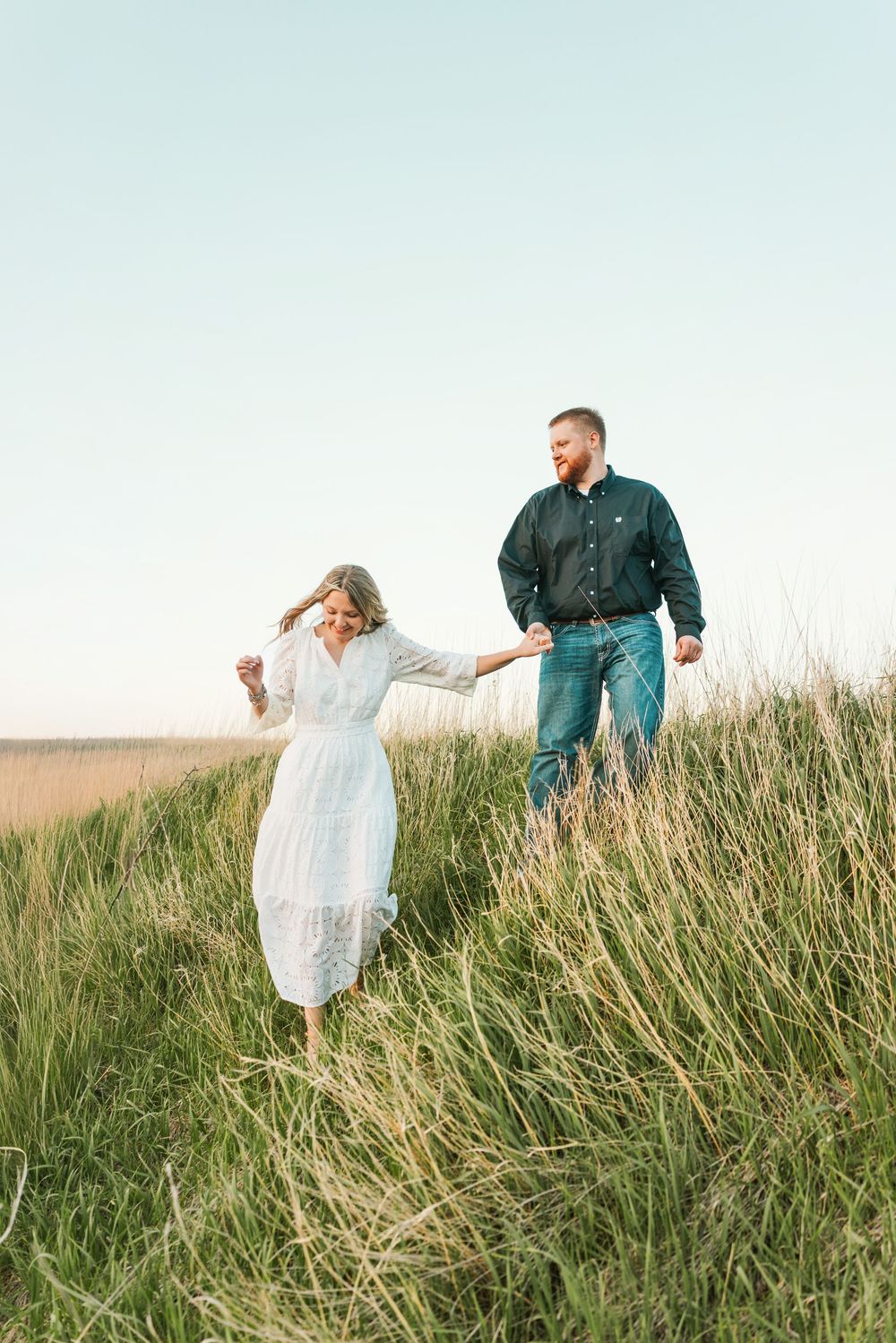 Couple walking hand in hand through tall green grass field, woman in white midi dress and man in dark shirt and jeans.