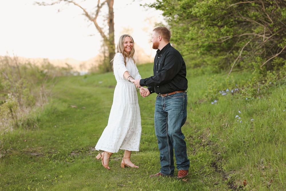 A couple holds hands and smiles on a lush green path, woman in white dress, man in black shirt and jeans.