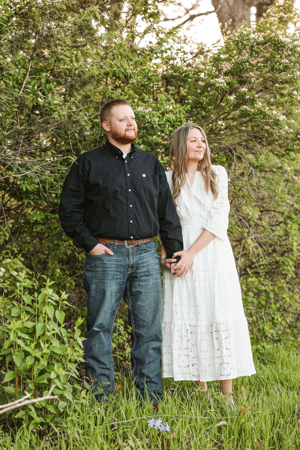 Couple holding hands standing in front of lush green bushes, woman in white dress and man in black shirt and jeans.